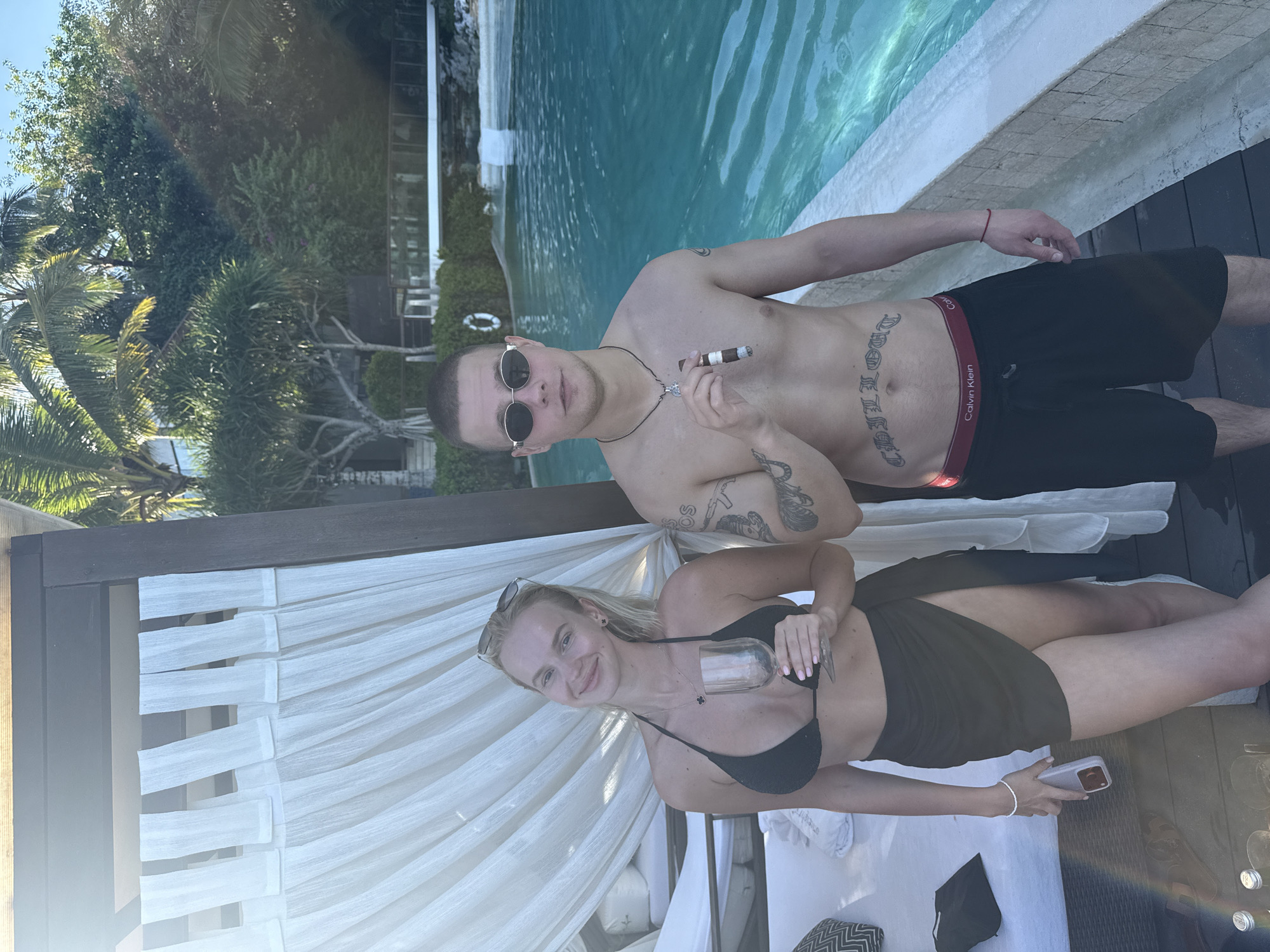 Two individuals are posing for a photo near a swimming pool in what appears to be a resort in Kuta Selatan, Indonesia. A man with short hair and sunglasses is shirtless, wearing black swim trunks with a red Calvin Klein waistband. He holds a cigar in his hand and has tattoos on his chest and arms. A woman in a black bikini and a black cover-up is holding a wine glass in her hand and a cell phone in the other. She has her blonde hair pulled back. They are both positioned near a white draped cabana. The background includes a view of the pool and surrounding vegetation, suggesting a tropical setting.