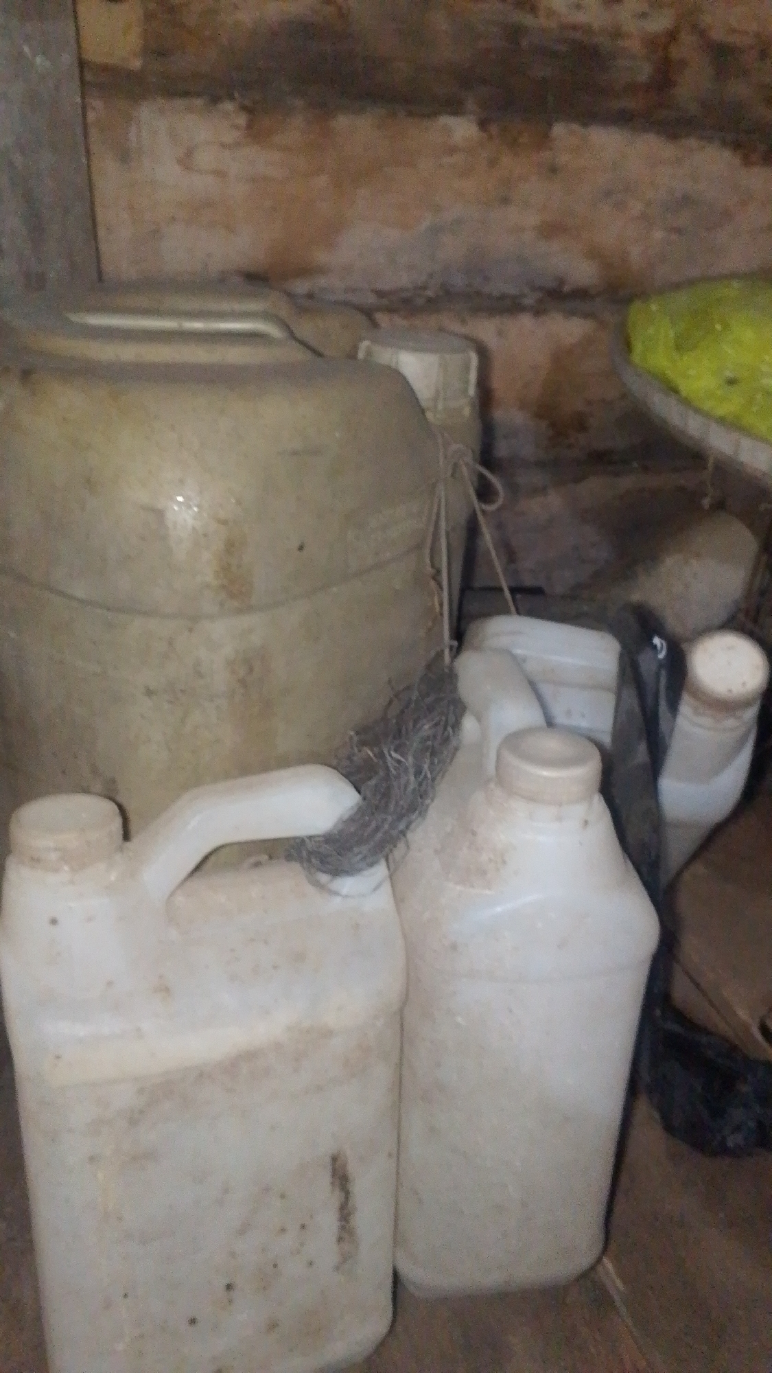 The image depicts a dimly lit storage area, likely a shed or utility room, in Olak-Olak Kubu, Indonesia. The scene is dominated by several large, white plastic containers of varying sizes, suggesting storage for liquids. In the foreground, a smaller jug with a handle and a cap is visible. Behind it, another similar jug is partially obscured. To the left, a very large, round, light-colored container stands prominently. A greyish, fibrous material, possibly nesting material or insulation, is bundled and tied to the spout of one of the smaller jugs. The walls appear to be rough and textured, with a reddish-brown hue, and show signs of dirt and age. A corner of a bright yellow object, possibly a tarp or plastic sheeting, is visible in the upper right portion of the image. The overall impression is one of disarray and storage of potentially useful but untidy items in a rural or utilitarian setting. There are no people present in the image.