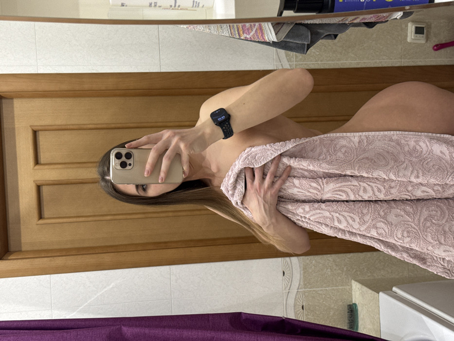 A woman stands in a bathroom in front of a mirror, holding a smartphone to take a selfie. The background shows a wooden door, white tile walls, and a portion of a bathroom sink. The woman is partially covered by a large, patterned pink towel, revealing a glimpse of her shoulder, hip, and upper thigh. She's wearing a smartwatch on her left wrist. Her hair is long and light brown.
