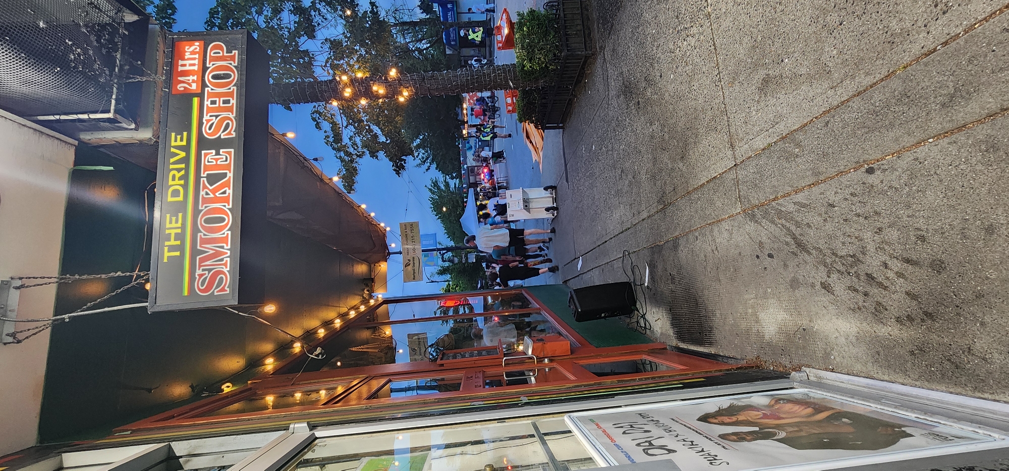 An eye-level, outdoor, medium shot shows a street scene at dusk in an urban environment. The foreground features a sidewalk with a few scattered leaves and a chain imbedded in the concrete. To the left, the exterior of a business is visible, with a glass door and windows reflecting the street. A large black speaker sits on the sidewalk near the entrance. Above the storefront, a black sign with yellow and red lettering reads "THE DRIVE SMOKE SHOP 24 Hrs." Small, lit bulbs hang from the awning, casting a warm glow.
In the midground, several people are walking on the sidewalk, some heading away from the camera. A person is pushing a wheeled cart. Further down the street, tents are set up, suggesting a market or event. Trees line the street, some decorated with strings of lights. The sky is a deep blue, indicating late evening. The overall atmosphere is casual and active.