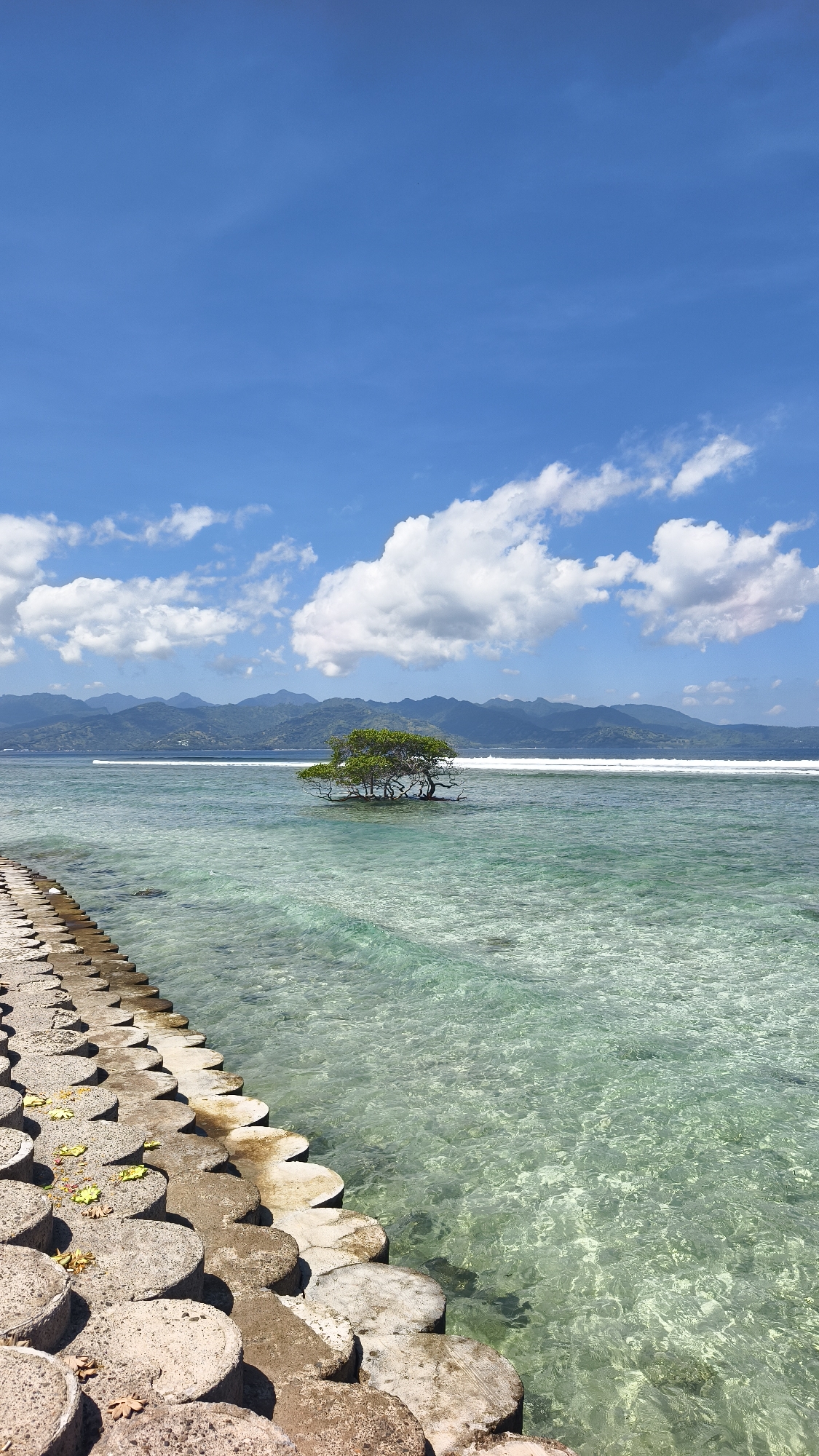 The image presents a scene at a tropical beach near the city of Unknown, Indonesia. The foreground consists of a man-made concrete structure designed for coastal protection. The clear, turquoise-colored water extends from the barrier toward a small islet in the middle ground. A solitary tree stands on the islet. In the background, mountains are visible along the horizon. The sky is bright blue with scattered white clouds.