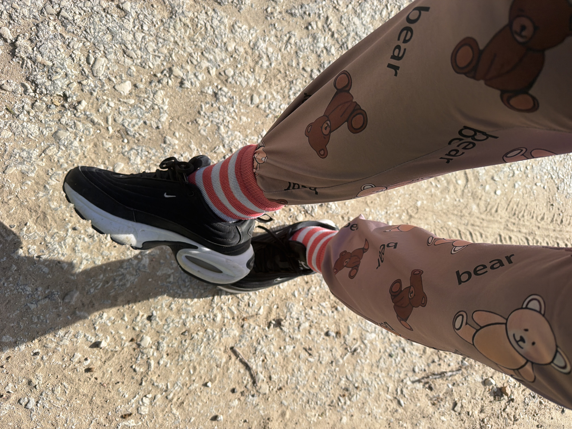 The image captures a top-down view of a person's lower legs and feet, likely standing or walking outdoors. The person is wearing black athletic shoes with prominent white soles, featuring a small white Nike swoosh visible on the side of one shoe. White socks with alternating pink or light red stripes are visible at the ankles.
The most distinctive clothing item is a pair of loose-fitting, light brown or tan pants. These pants are adorned with a repeating pattern of cartoon-style brown teddy bears in various poses, interspersed with the word "bear" printed in black lowercase letters.
The setting is an outdoor, unpaved area. The ground consists of light-colored sand and small, off-white gravel or pebbles, suggesting a dirt path or a natural, open space. Strong, defined shadows are cast on the ground, indicating bright sunshine and a clear daytime sky, possibly morning or late afternoon given the shadow length. The scene takes place in Priseltsi, Bulgaria.