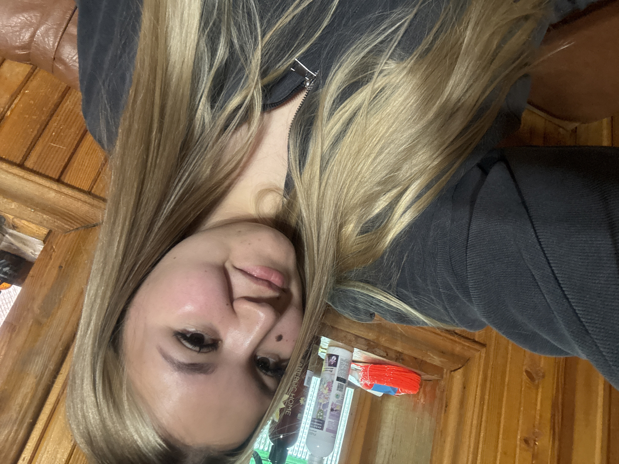 A young woman with long, blonde and light brown hair is featured in a close-up, sideways-oriented selfie. She is wearing a dark, possibly grey or black, zip-up top or hoodie, visible around her shoulders and chest. Her head is slightly tilted, and she displays a soft, relaxed smile. Key facial details include dark eyes with noticeable eyelashes, defined eyebrows, and a small mole on her left cheek (viewer's right).
The background indicates an indoor setting, characterized by warm, reddish-brown wooden paneling or furniture. In the upper left corner, a section of a brown, possibly leather, surface is visible. On a surface in the lower right, various household items are present, including several bottles and a red and blue brush-like object. One bottle clearly displays the text "FABERLIC HOME" along its side. Another bottle next to it features a floral design and appears to have text in a foreign language. The image conveys a personal and casual moment, likely taken within a home in Большой Букор, Russia.