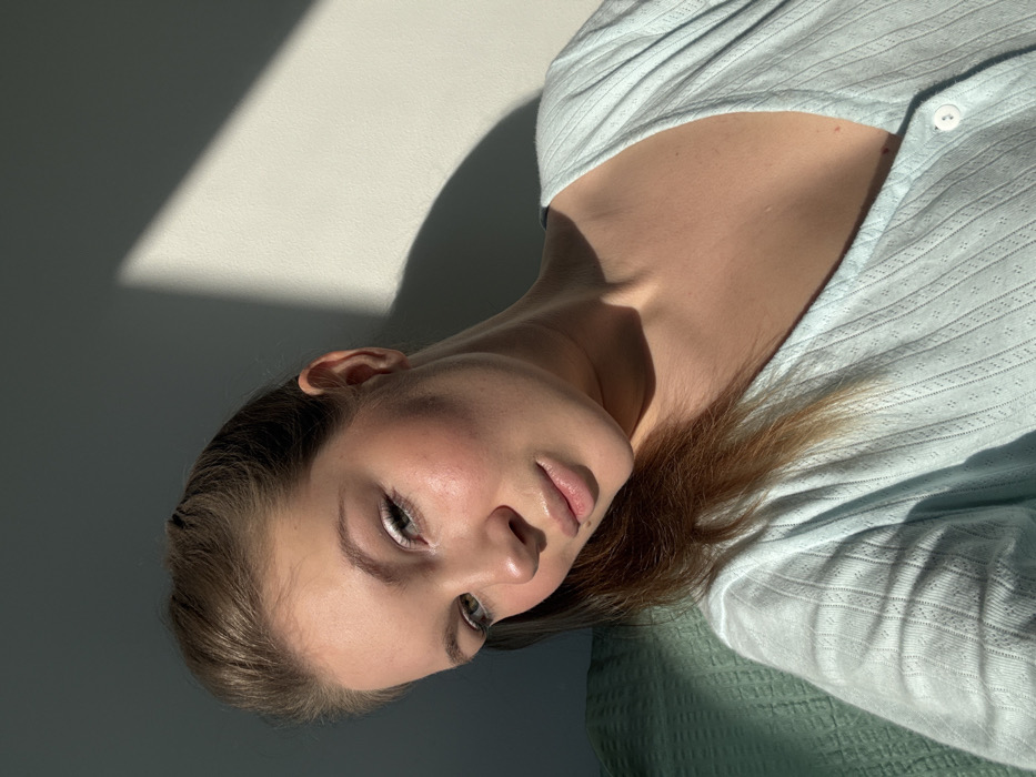 A young adult female is depicted in a close-up, indoor selfie, located within the city of Vozdovac Urban Municipality, Serbia. She has light skin, light brown hair styled upwards from her forehead, and light-colored eyes. Her face shows subtle makeup, including white eyeliner on the lower lash line, blush on her cheeks, and glossy lips. She is wearing a light blue, V-neck garment with a textured pattern and a visible white button on the front placket. Her head is slightly tilted, and she looks directly at the camera with a neutral expression. Direct sunlight illuminates her face and upper body, casting a distinct triangular shadow on the plain, light-colored wall behind her to the right. A textured, teal or mint green fabric is visible in the lower left portion of the frame, behind her.