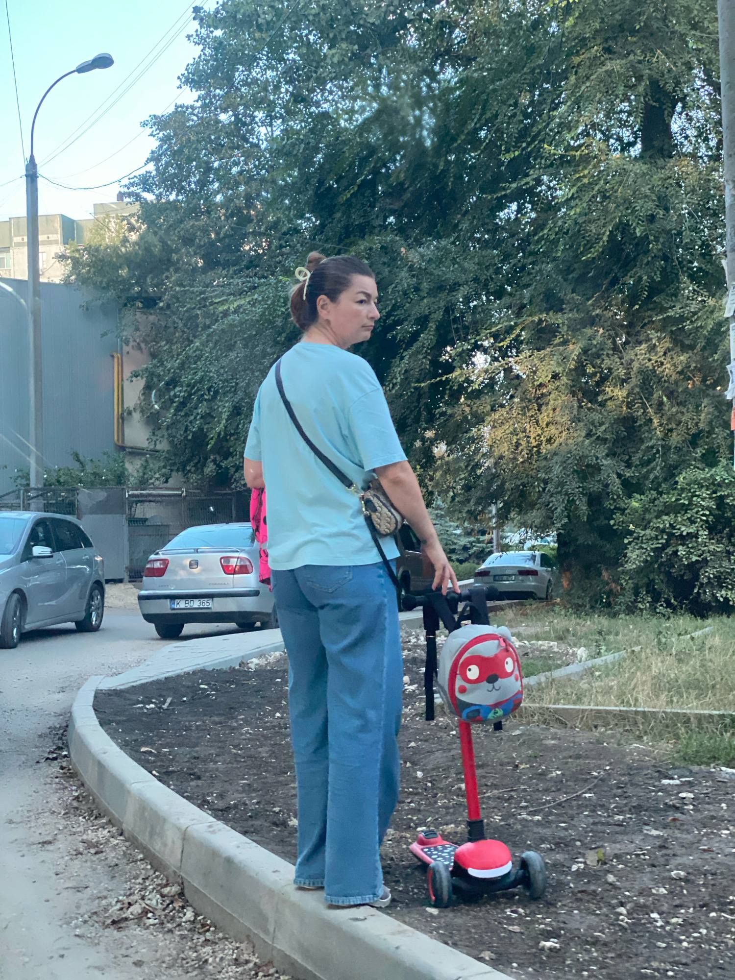 A woman stands on a sidewalk next to a red scooter with a child's helmet attached to the handlebars. The helmet has a cartoon character design. The woman is wearing a light blue t-shirt and blue jeans. She has a crossbody bag with a leopard print. The scooter is positioned on a patch of dirt and gravel. In the background, there are two cars parked on the street, one silver and one grey. A street lamp is visible to the left, and trees and bushes line the background. The scene appears to be in a residential area of Chișinău, Moldova, possibly near an apartment building based on the structure visible behind the trees on the left. The license plate on the silver car reads "K BD 365". The overall lighting suggests it is daytime, likely late afternoon due to the quality of the light.