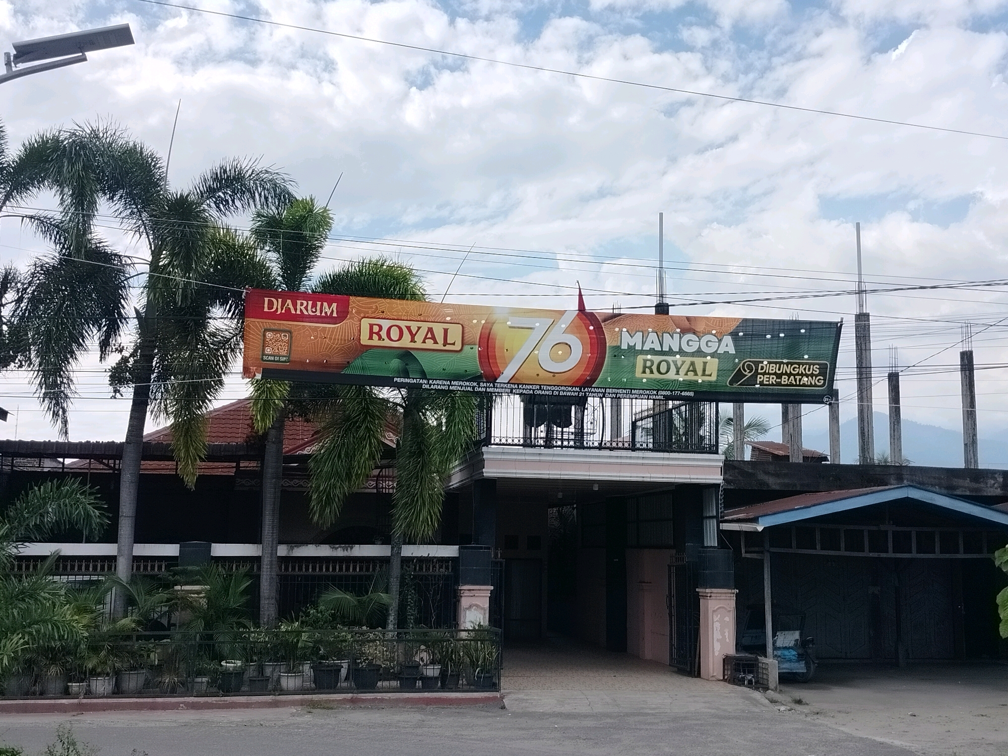 A street-level, eye-level, wide-angle shot captures a billboard for Djarum Royal 76 Mangga Royal. The billboard is a banner stretched across the front of a building, featuring product branding and warning messages about smoking. The banner's colors are vibrant, with oranges, reds, and greens dominating the design. The building itself appears to be a commercial establishment, possibly a storefront or office, with a dark-colored facade and a covered entrance. Several palm trees are visible on the left side of the image, their fronds reaching towards the sky. The sky is partly cloudy, with a mix of white clouds and blue sky. The street in front of the building is paved and appears to be relatively clean. A few potted plants are lined up along a low fence in front of the building. The scene is set in a tropical or subtropical environment, consistent with the location of Pulonasbaru, Indonesia. There are no people visible in the image.