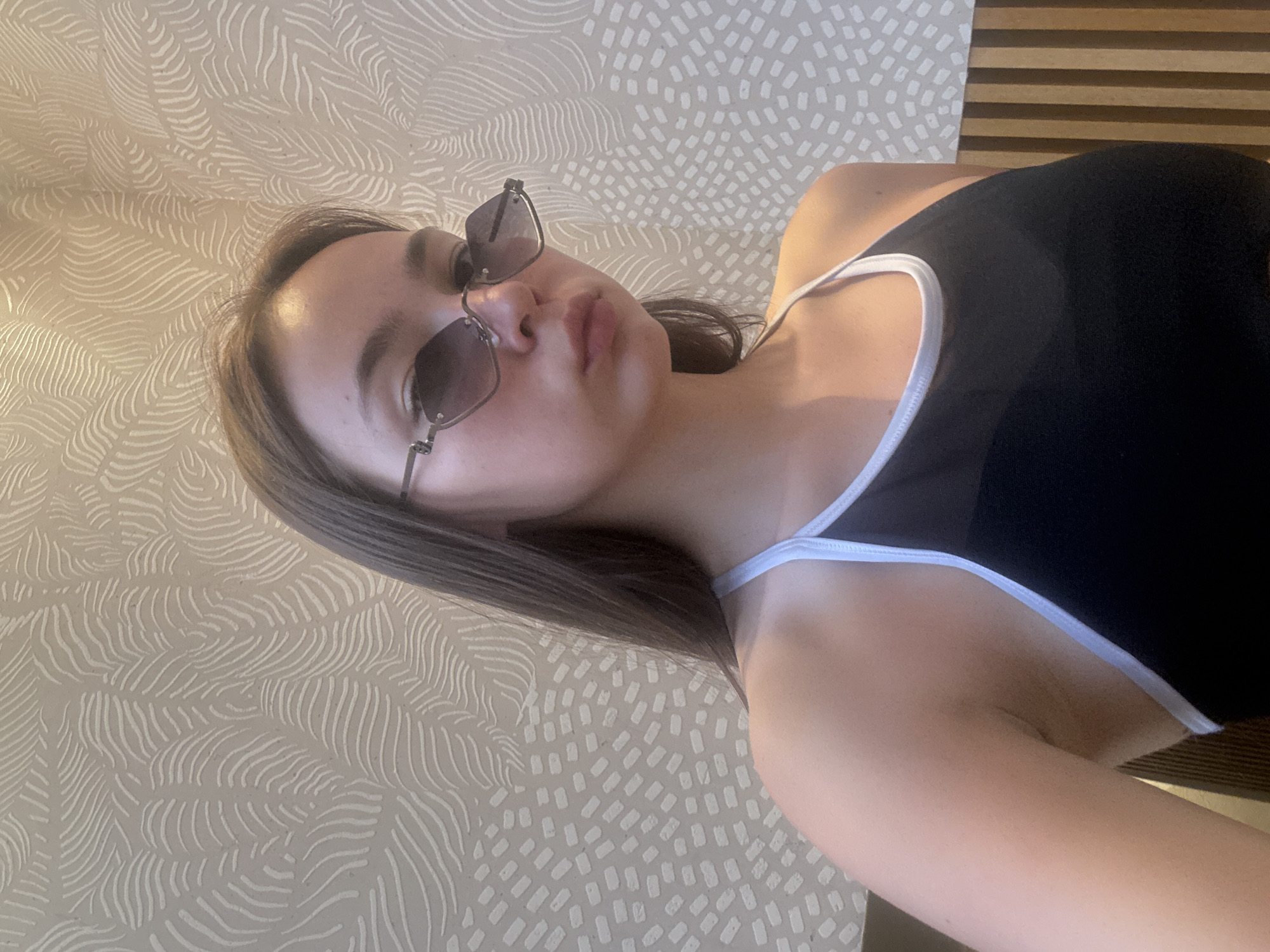 A woman is posing for a selfie indoors. She wears sunglasses and a black tank top with a white trim. The background is a patterned wall.