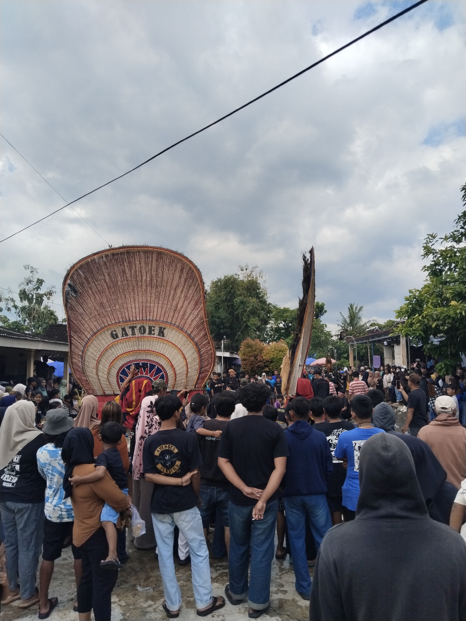 A vibrant outdoor cultural event is captured in Mojodoyong, Indonesia, with a large crowd gathered under an overcast sky. The focal point of the scene is a massive, ornate, fan-shaped structure in the center-left, made from woven natural materials, possibly bamboo or reeds. The word "GATOEK" is prominently displayed in large letters across its lower section. This structure appears to be part of a float or a traditional performance prop. To its right, another tall, dark, abstract wooden or natural material structure stands upright, adding to the ceremonial feel.
The event is bustling with activity, as numerous people of all ages, including children, are packed closely together, intently watching the unfolding spectacle. Most individuals are seen from behind, indicating they are facing the central performance area. Simple, single-story buildings with corrugated roofs and lush green trees form the backdrop, suggesting a rural or semi-urban community setting. Overhead power lines crisscross the sky.
The weather is cloudy but bright, indicating it is daytime, likely late morning or afternoon. The ground appears to be a mix of dirt and possibly damp areas from previous rain or activity. People are dressed in casual attire, with some wearing traditional head coverings. Visible text includes "GATOEK" on the large prop, and on a black t-shirt in the foreground, "AVENGED SEVENFOLD" is legible, with "SEDIA" on another shirt to the far left. The overall atmosphere suggests an engaged community participating in a local cultural festival or parade.