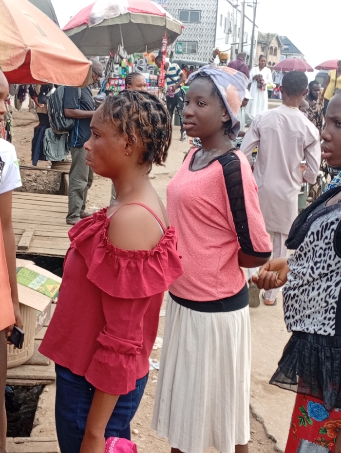 The image depicts a bustling outdoor market scene in Ura Nta Umuarandu, Nigeria. In the immediate foreground, three females are partially visible. On the far left, a person in a light-colored top is partially cropped. Centrally positioned, a young adult female with dark braided hair is seen in left profile, wearing a dark red off-the-shoulder top with ruffled sleeves and dark blue trousers. She stands on a wooden plank walkway that spans an open drainage ditch containing refuse, with a cardboard box nearby. To her right, another young adult female faces right in profile, wearing a light pink and black short-sleeved top, a pale patterned headscarf, and a long pleated cream-colored skirt; a patch of white residue is visible on her neck. On the far right, a third female, partially cropped, wears a black and white patterned top over a black ruffled skirt and a red floral-patterned skirt.
The midground is a crowded street filled with numerous individuals. A man with a dark backpack is visible behind the woman in red. Farther back, a person in a light-colored traditional Nigerian garment is prominent. Market stalls displaying various goods, including electronics, are set up under large umbrellas, one featuring red and white stripes. The background reveals multi-story buildings, some with distinctive patterned facades, and overhead power lines. The ground surface appears unpaved or uneven, consistent with an active urban market environment.