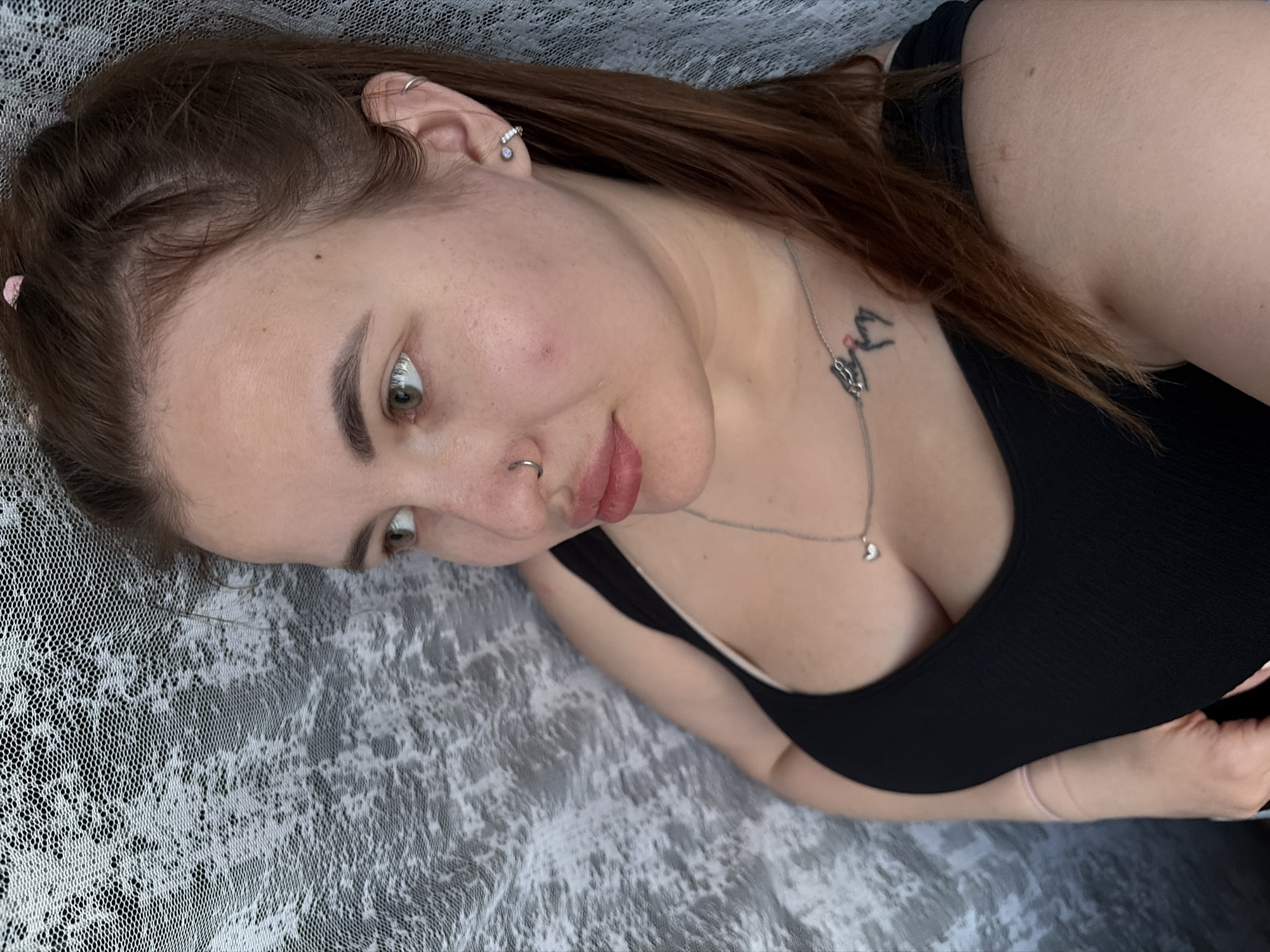 A woman is lying on a white and gray patterned surface, facing the camera at an angle. She has brown hair with a pink clip, light skin, and green eyes. She is wearing a black tank top and a silver necklace with a heart pendant. She has piercings in her ear and a nose ring. There is a tattoo visible on her chest. The location and city are unknown.