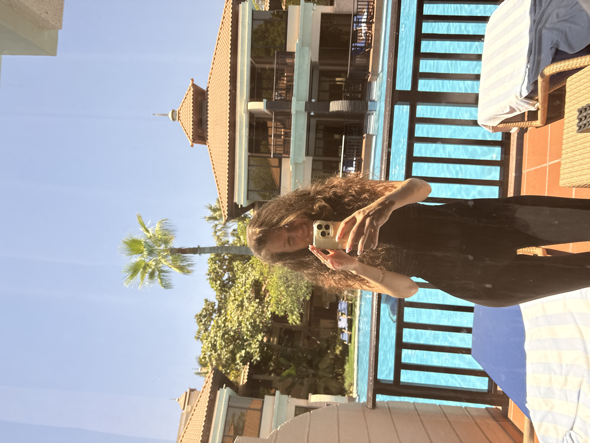 In this horizontal, natural light image, a woman with long, curly brown hair is captured in a selfie taken from the reflection in a glass balcony door. She is wearing a black, form-fitting outfit and holding a smartphone in her right hand, with her left hand near her face.
Behind her, reflected in the glass, is a resort-like setting. A vibrant blue swimming pool stretches across the background, bordered by a wooden railing. Beyond the pool, a multi-story building with terracotta-tiled roofs and dark-framed windows is visible. Lush green trees and a tall palm tree are interspersed with the architecture, suggesting a warm climate. The sky is clear and bright blue. The balcony itself is paved with terracotta tiles and features lounge chairs with blue and striped towels.
The scene appears to be a hotel or resort located in Dubai, United Arab Emirates, characterized by its upscale architecture, clear blue skies, and resort amenities.