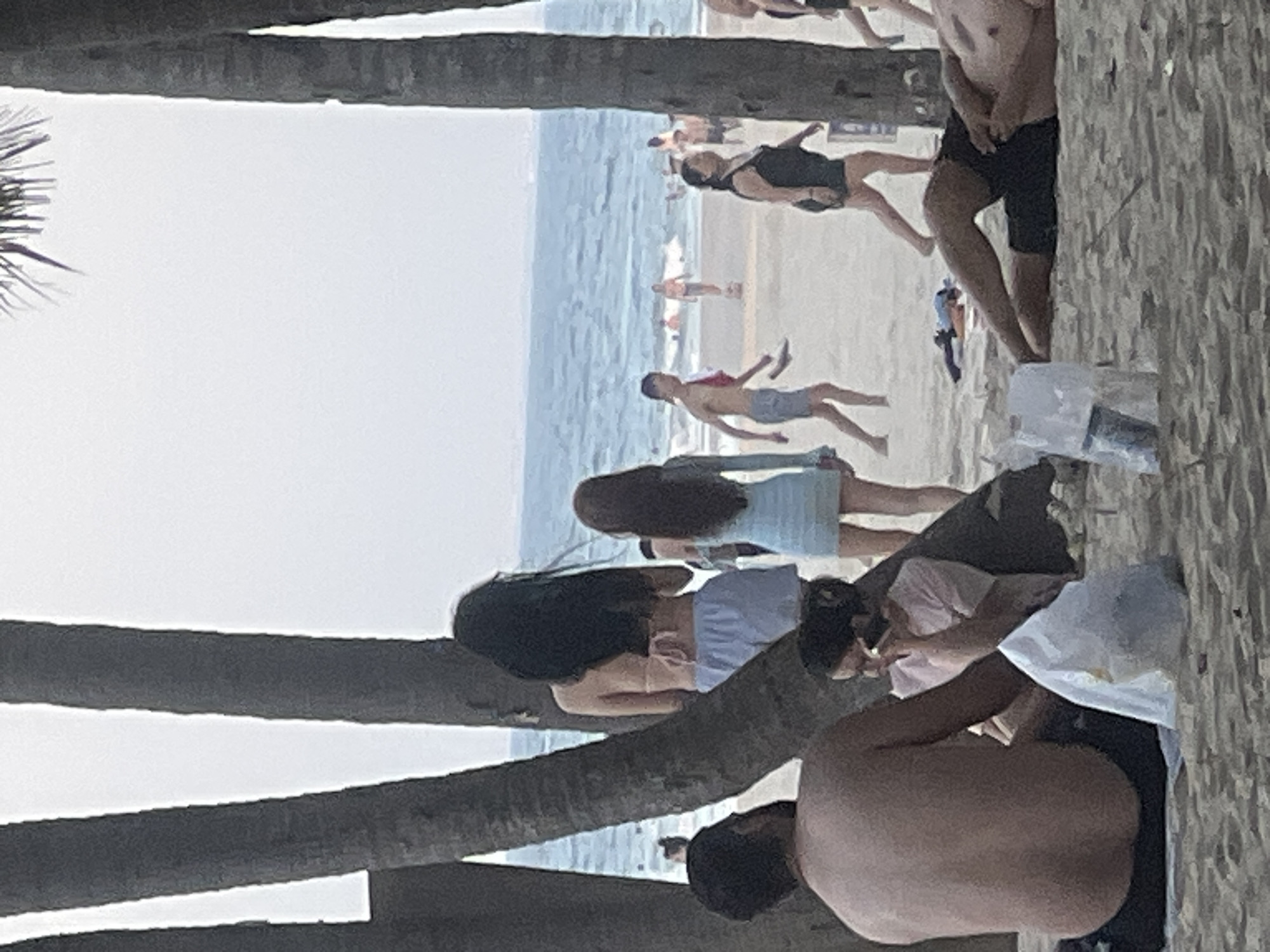 The image depicts a scene on a beach, likely in Đà Nẵng, Vietnam, with the ocean and sky visible in the background. Several palm trees are present, framing the view. In the foreground, a man is seated with his back to the camera, wearing dark shorts. Next to him, a woman is also seated, wearing a light-colored top and holding a phone to her ear. A white plastic bag sits between them.
Further back on the sandy beach, two women are walking away from the camera. One is wearing a light blue dress, and the other is wearing a light-colored bikini top and light blue shorts. Several other people are visible in the mid-ground, some walking and some in the water. A man in dark shorts is lying on the sand in the lower right portion of the image. The overall atmosphere is that of a casual beach day.