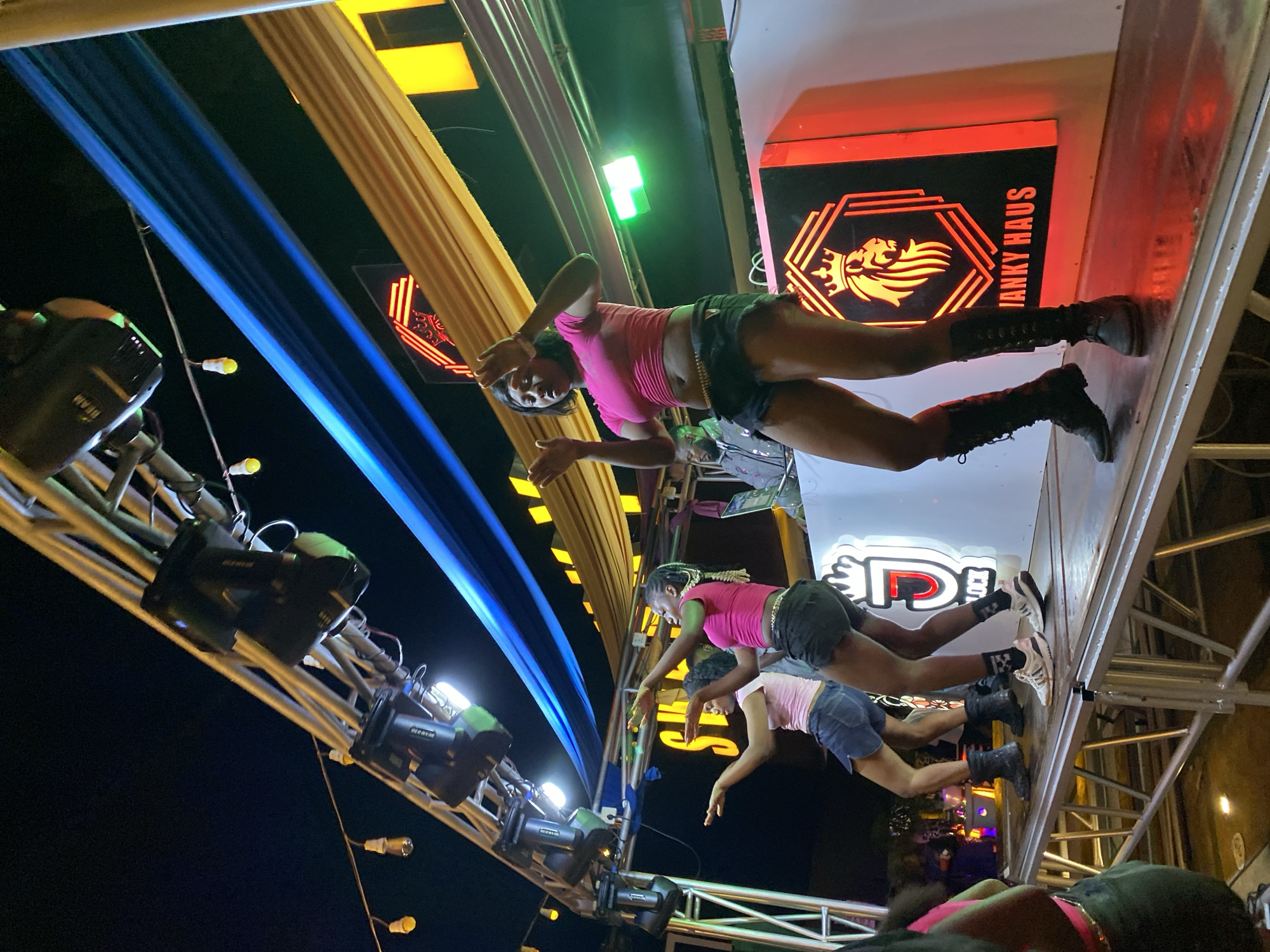 In Jos, Nigeria, a group of young women is performing on a stage at night. The stage is elevated and made of metal, with lights and fabric draping from above, some of which are blue and yellow. The women are dancing energetically, with one woman in the foreground wearing a pink top, black shorts, and black boots. She has a beaded belt around her waist. Another woman next to her wears a pink top and a denim skirt, with white sneakers. Behind them, another performer in a pink top and a denim skirt is visible. A DJ booth with a logo featuring a lion and the text "JANKY HAUS" is set up on the stage. The background shows a crowd of people and more lighting. The overall atmosphere is that of a lively outdoor event.