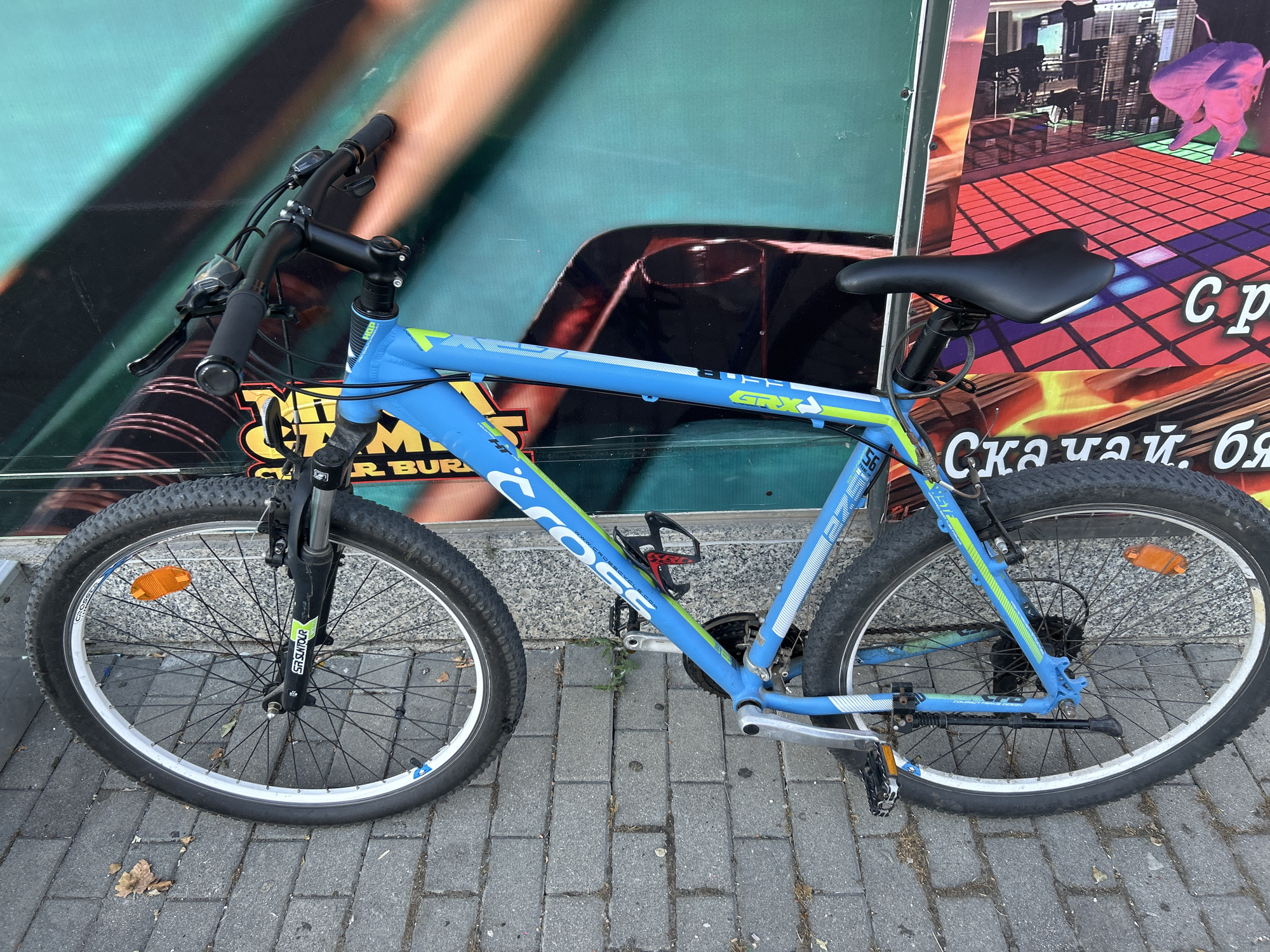 The image shows a blue and green mountain bike parked on a paved sidewalk in front of a building with a large, colorful mural. The mural features abstract shapes and figures, including what appears to be a hand and city buildings. The bike has black handlebars with grips and shifters, a black seat, and black tires with silver rims. The front fork is black with the "SR SUNTOUR" logo visible. The bike's frame has the brand name "CROSS" printed in large white letters. There is a water bottle cage attached to the frame. The sidewalk is made of gray rectangular pavers, and some dry leaves are scattered on the ground. The lighting suggests it is daytime. The scene is consistent with an urban environment, possibly in the city of Burgas, Bulgaria, given the presence of Cyrillic text on the mural which appears to say "Скачай, бя" (Download, bя). There are no people visible in the image.