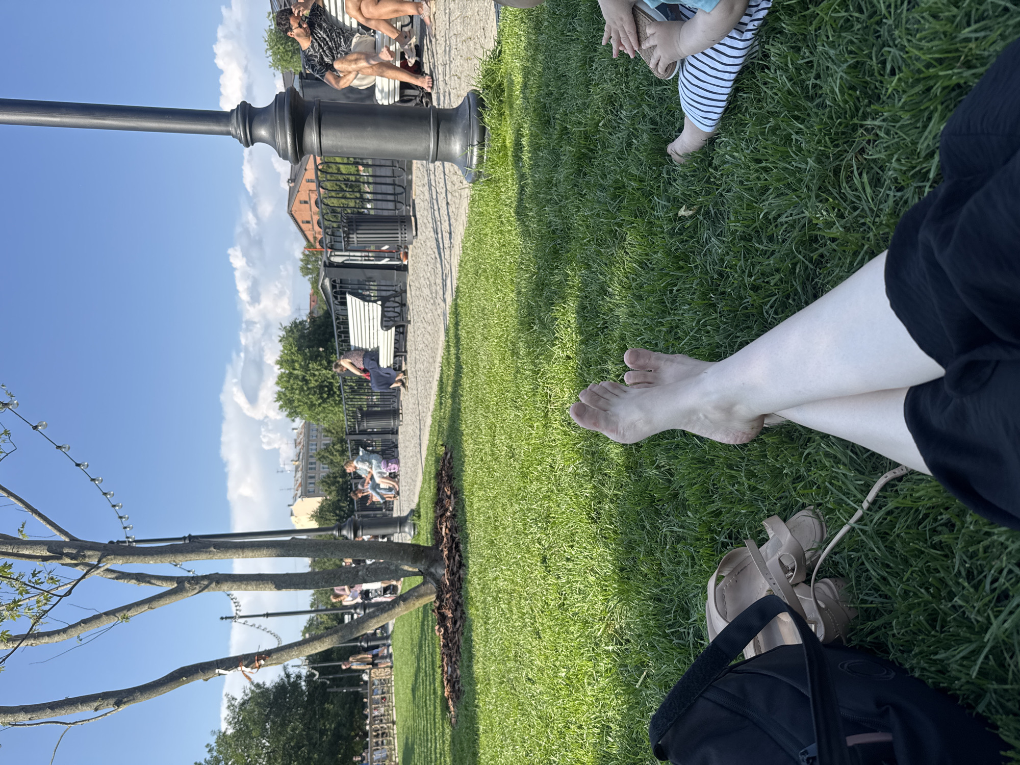 The image shows a group of people relaxing on a grassy area in what appears to be a park or public space. The perspective is from a person sitting on the grass, looking towards a path and a row of benches with more people seated.
In the foreground, the legs and feet of the person taking the photo are visible, along with a pair of sandals and a black bag. The grass is lush and green, with some darker mulch around the base of a tree.
In the middle ground, a paved path runs alongside the grass, bordered by a black metal fence. Several people are sitting on white benches along the path, some talking and others looking around. A large, dark lamppost stands near the path. Beyond the path, buildings with brick facades and windows are visible, suggesting an urban setting.
In the background, the sky is blue with scattered white clouds, indicating a clear, sunny day. Trees with green foliage are scattered throughout the scene. The overall impression is one of a relaxed outdoor gathering on a pleasant day. The time of day appears to be daytime, possibly late morning or afternoon, given the bright sunlight and clear skies. There is no visible text in the image.