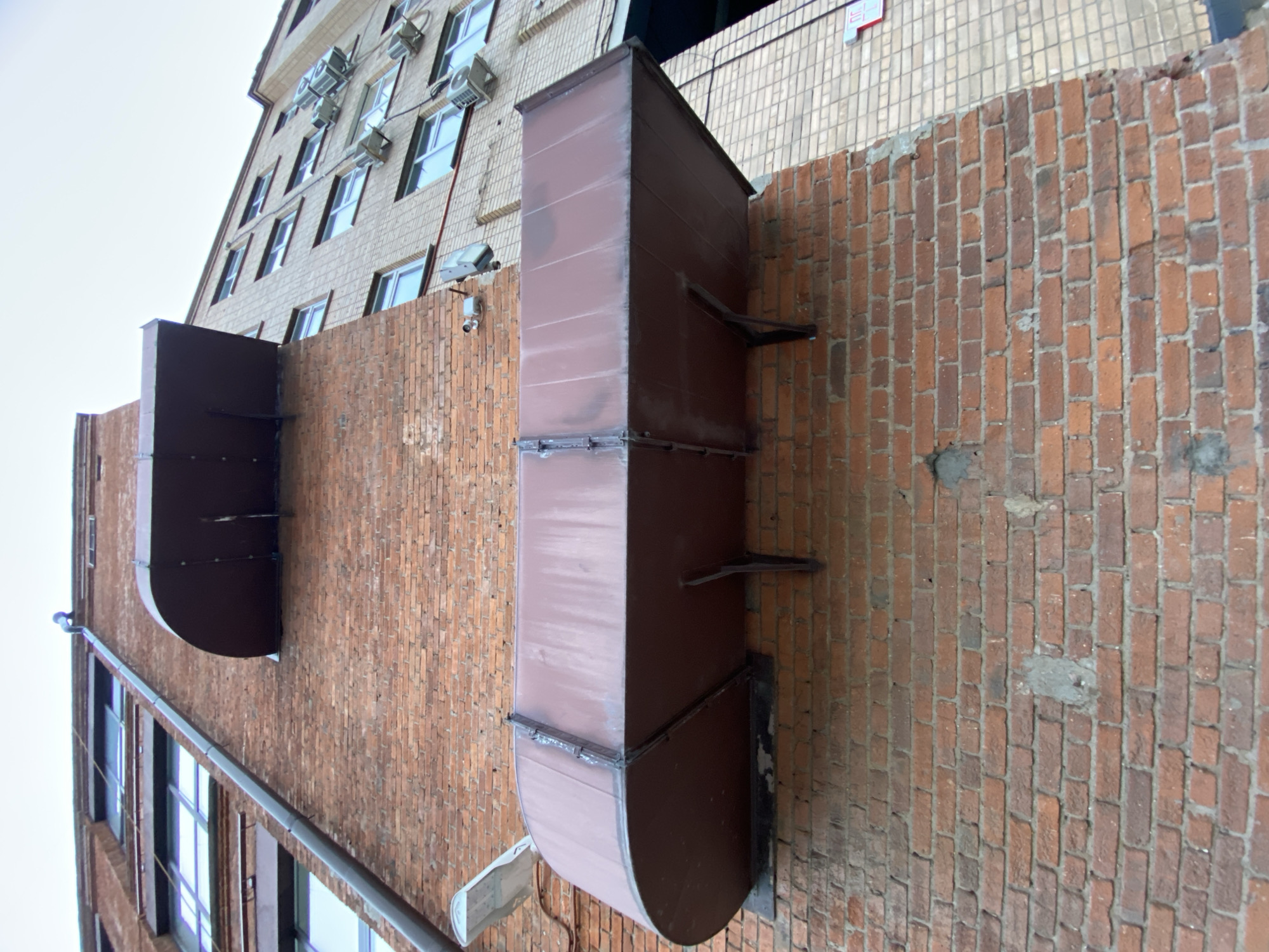 The image displays the exterior of a multi-story building in Moscow, Russia, observed from a low angle under an overcast daytime sky. The building's side wall is prominently featured, segmented into two distinct material sections: a lower part constructed from aged red bricks with visible mortar, and an upper part featuring a lighter-colored brick or tiled facade.
Two large, rectangular, dark brown metal ventilation ducts are central to the composition. One longer duct runs vertically, extending from the lower portion of the red brick wall upwards. Another shorter, curved duct is situated to its left, also mounted on the red brickwork. Both ducts are secured to the wall with metal brackets.
On the upper, lighter-colored section of the building, numerous rectangular windows with light blue reflections are visible, each accompanied by small outdoor air conditioning units mounted beneath them. Several small white security cameras and lighting fixtures are affixed to the red brick wall. A dark-colored horizontal drainpipe runs along the top edge of the red brick section, extending towards the left.
There are no people or specific activities taking place. The overall impression is one of an urban architectural scene. A small, red rectangular sign with indistinct white text is faintly visible on the upper part of the building.