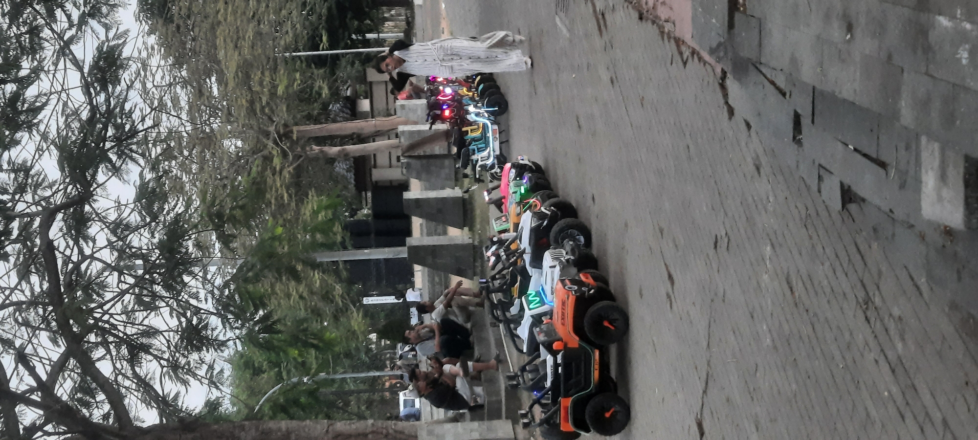 A line of colorful toy electric cars, resembling jeeps and scooters, is parked on a paved walkway. A woman wearing a striped outfit walks away from the camera, passing by the toy vehicles. In the background, several people are seated on steps, observing the scene. Trees with dense foliage provide shade over the area. The setting appears to be an outdoor public space, possibly a park or recreational area in Kanigoro, Indonesia.