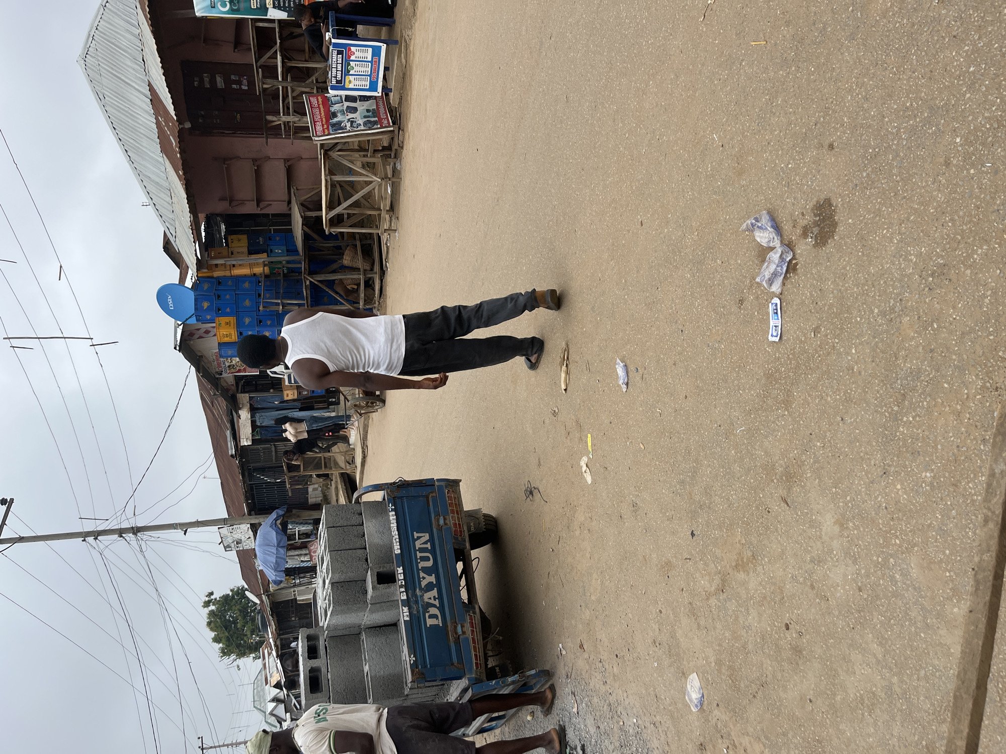 This image depicts a street scene in Abuja, Nigeria, likely a market area. A blue motorized tricycle, labeled "DAYUN", is parked on the left side of the frame, loaded with stacked concrete blocks. Two men are visible, one wearing a green cap and a white t-shirt with green lettering, standing near the tricycle. Another man, wearing a white tank top and dark trousers, is walking away from the tricycle towards the right side of the image. The street is unpaved and dusty, with scattered debris and wrappers on the ground. A row of shops with corrugated metal roofs lines the right side of the street, some appearing to be open with goods displayed, while others appear closed or under construction. A blue satellite dish is mounted on one of the shop roofs, along with numerous electrical wires crisscrossing overhead. The sky is overcast.
