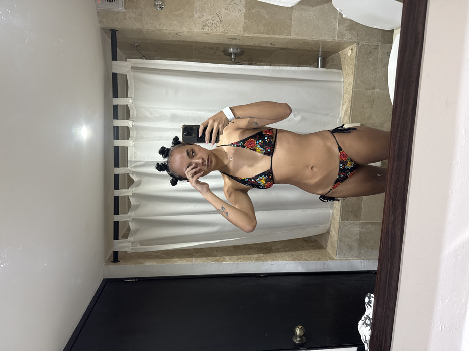 The image depicts a young adult female reflected in a mirror, positioned indoors, likely within a private bathroom of a hotel or residence in Taxco, Mexico. The specific address or public location within Taxco is not discernible.
The subject is a woman with dark hair styled in multiple small knots or buns. She is wearing a two-piece bikini featuring a black base with a colorful floral pattern of red, yellow, and blue flowers with green foliage. A small, delicate, gold-colored necklace is visible around her neck. She has tattoos on both arms, including a small butterfly-like design on her left bicep. On her right wrist, she wears a white smartwatch. Her left hand is raised to her left eye, with fingers touching the eye area, while her right hand holds a dark-colored smartphone, capturing the mirror selfie. Her expression is neutral to slightly fatigued.
The background, reflected in the mirror, shows a bathroom environment. To the left, a dark-colored door with a metallic doorknob is visible. The wall behind the subject and to the right is tiled with light-colored, possibly travertine, stone. A white shower curtain hangs on a dark rod across the width of a shower stall. Shower fixtures are partially visible on the right tiled wall. Above, a white ceiling with a single recessed light fixture is present, along with a white exhaust fan located on the upper right wall. A white toilet bowl is partially visible on the far right, and a white towel hangs on a rack. At the bottom edge of the image, a dark wooden frame, likely part of the mirror's structure, reflects a crumpled garment with a white and black pattern on the left.