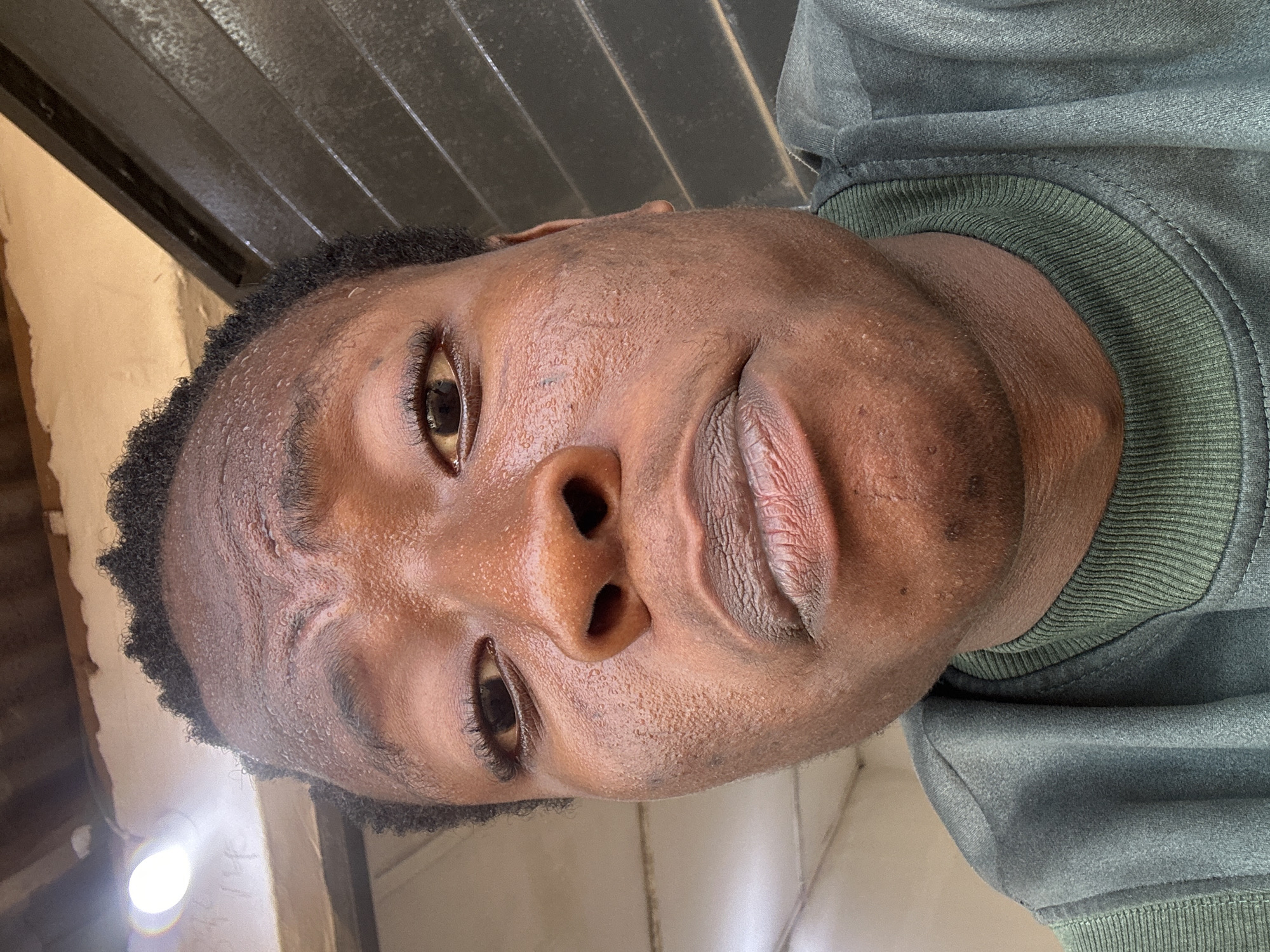 A close-up, angled portrait shows a young Black man looking down towards the camera. The man has short, dark, curly hair. His skin appears damp or sweaty. He is wearing a dark green, possibly sweatshirt-like garment. The background includes dark, corrugated material. At the bottom left, a faint light source is visible. The location is unknown.