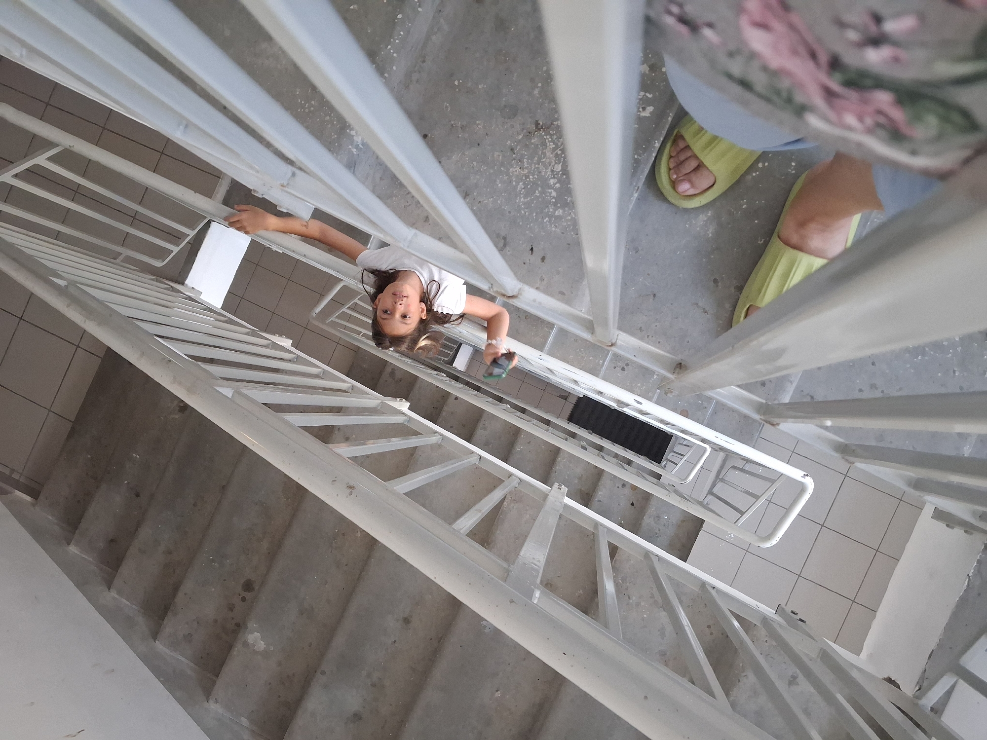 In a concrete stairwell with a white metal railing, a young girl with brown hair, wearing a white t-shirt, is looking up at the camera and holding a green object in her right hand. She is positioned on a staircase that angles upwards and to the right. Below her, a pair of light green slides are visible on a concrete step, and a floral-patterned fabric is also partially visible. The stairwell appears to be located in Novosibirsk, Russia.