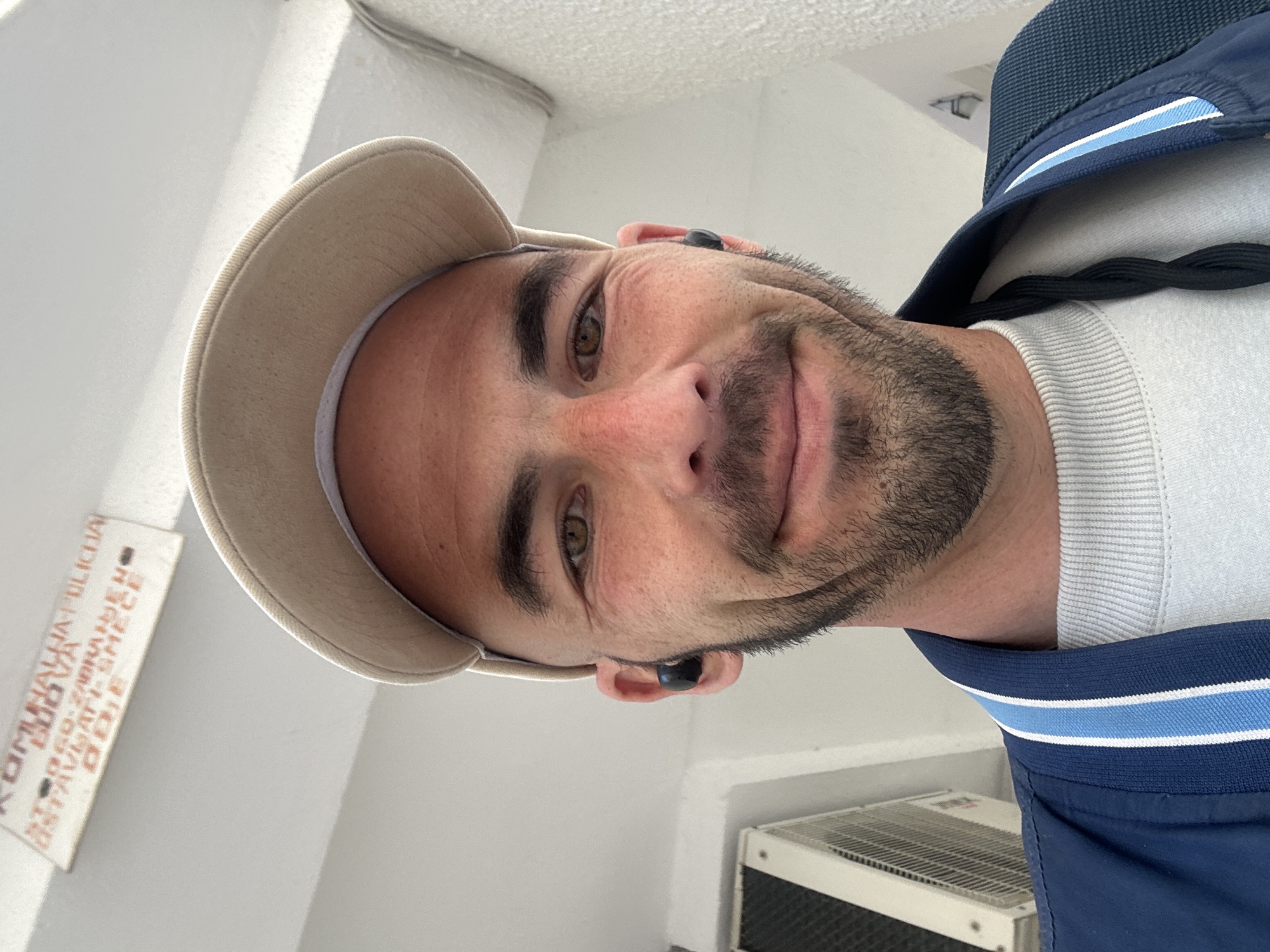 A selfie of a man with light brown eyes, dark eyebrows, a beard, and short dark hair. He is wearing a beige baseball cap, a white crewneck sweater, and a dark blue jacket with light blue stripes around the collar. He has a black wireless earbud in each ear. The photograph is taken indoors against a white wall. A sign with illegible text is to the left of the man. A white air conditioning unit is visible in the lower right corner of the image. Based on the sign visible in the image, it is likely the picture was taken in Budva, Montenegro.