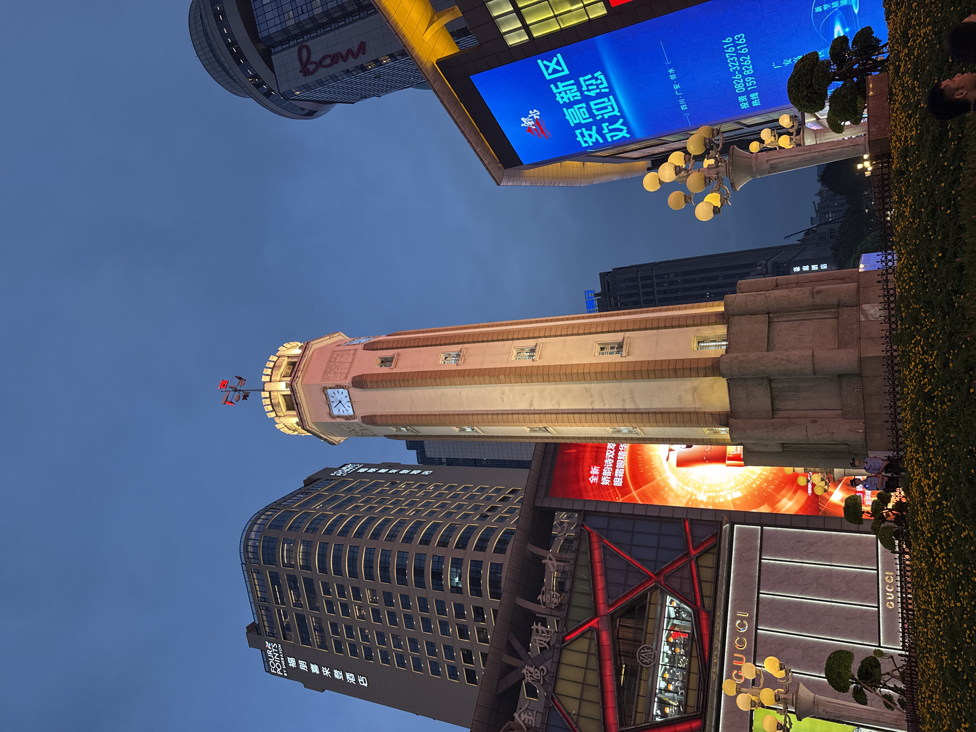 The image depicts an outdoor scene at dusk, likely in a commercial district of Yuzhong District, China, given the prominent signage. A clock tower, illuminated by warm light, stands centrally, topped with a weather vane displaying red flags. Surrounding the clock tower are modern buildings, including a skyscraper with "FOUR POINTS BY SHERATON" signage and another rounded building with the "Bani" logo visible in red. In the foreground, a well-maintained garden with yellow flowers and manicured bushes is bordered by a black metal fence. Several streetlights with multiple globes provide additional illumination. Adjacent to the clock tower, a large, bright blue digital billboard displays Chinese text, which appears to be a welcome message. To the left, a building with a red, geometric facade features large "GUCCI" logos. Two uniformed security guards are visible standing near the fence in the lower right portion of the image. The sky is a deep twilight blue, indicating the transition from day to night.
