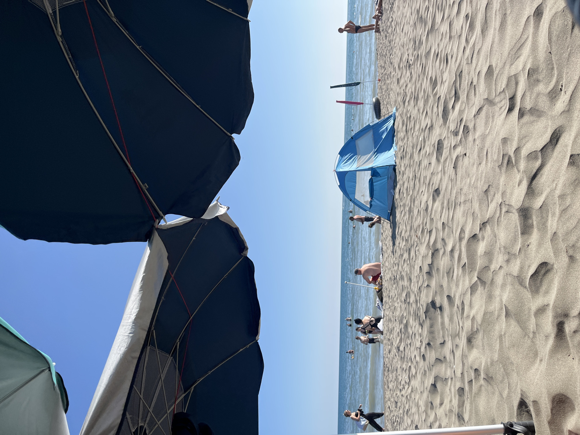 This is a photograph of a beach in Donji Štoj, Montenegro, taken from a low angle, looking across the sand towards the sea. In the foreground, there are large beach umbrellas, one dark blue and one partially visible green and white. The sand is textured with wind-blown ripples.
In the midground, several people are visible on the beach and in the shallow water. A small, blue pop-up tent is set up on the sand. There are also two colorful flags on poles, one pink and one green, and a black inflatable ring. Some individuals are wading in the water, while others are sitting or standing on the sand. One person is seen sunbathing.
The background consists of the calm, blue sea under a clear, bright blue sky. The horizon is clearly defined. The overall scene depicts a typical summer day at the beach with recreational activities.