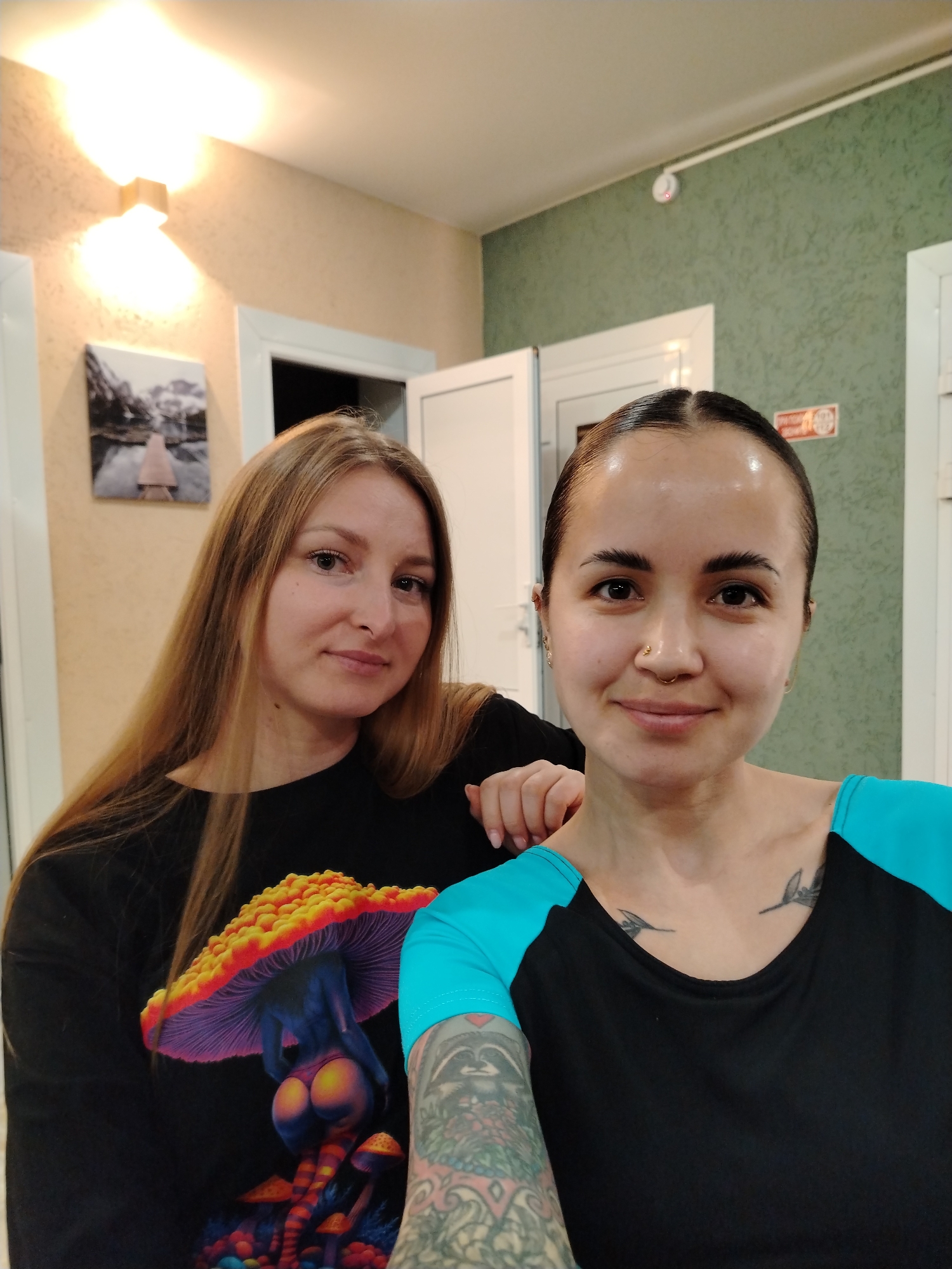 Two women pose indoors. The woman on the left has long, straight blonde hair and wears a black shirt with a graphic of a mushroom with human buttocks. The woman on the right has dark hair pulled back, a nose ring, a tattoo on her chest of two birds in flight, and a black and teal shirt. She also has a tattoo on her left arm. The location may be in Chaikovsky, Russia as indicated, but there is no other information in the image to verify this. The background is a cream-colored wall with a painting and a light fixture to the left, and a pale green wall with a fire safety sign and white door to the right. The lighting appears to be artificial.