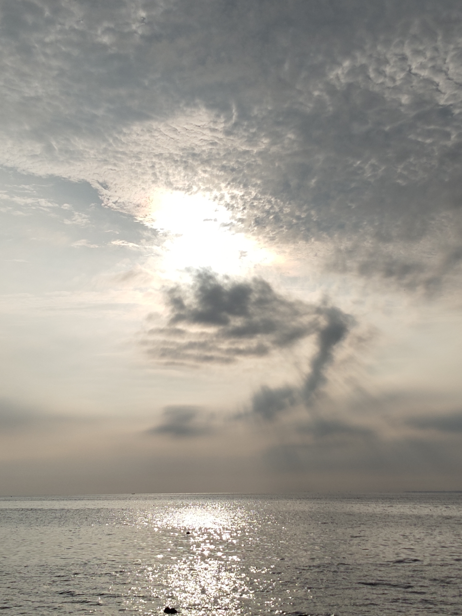 The image depicts a seascape at Lhokseumawe, Indonesia, characterized by a tranquil sea reflecting the sunlight. The sky is dominated by a layer of altocumulus clouds, with the sun obscured but its light diffusing through a break in the cloud cover. The sea surface is rippled, creating a specular reflection of the sun. The photograph is taken during daylight hours, with the light source appearing to be in a high position. The water appears calm with slight waves.