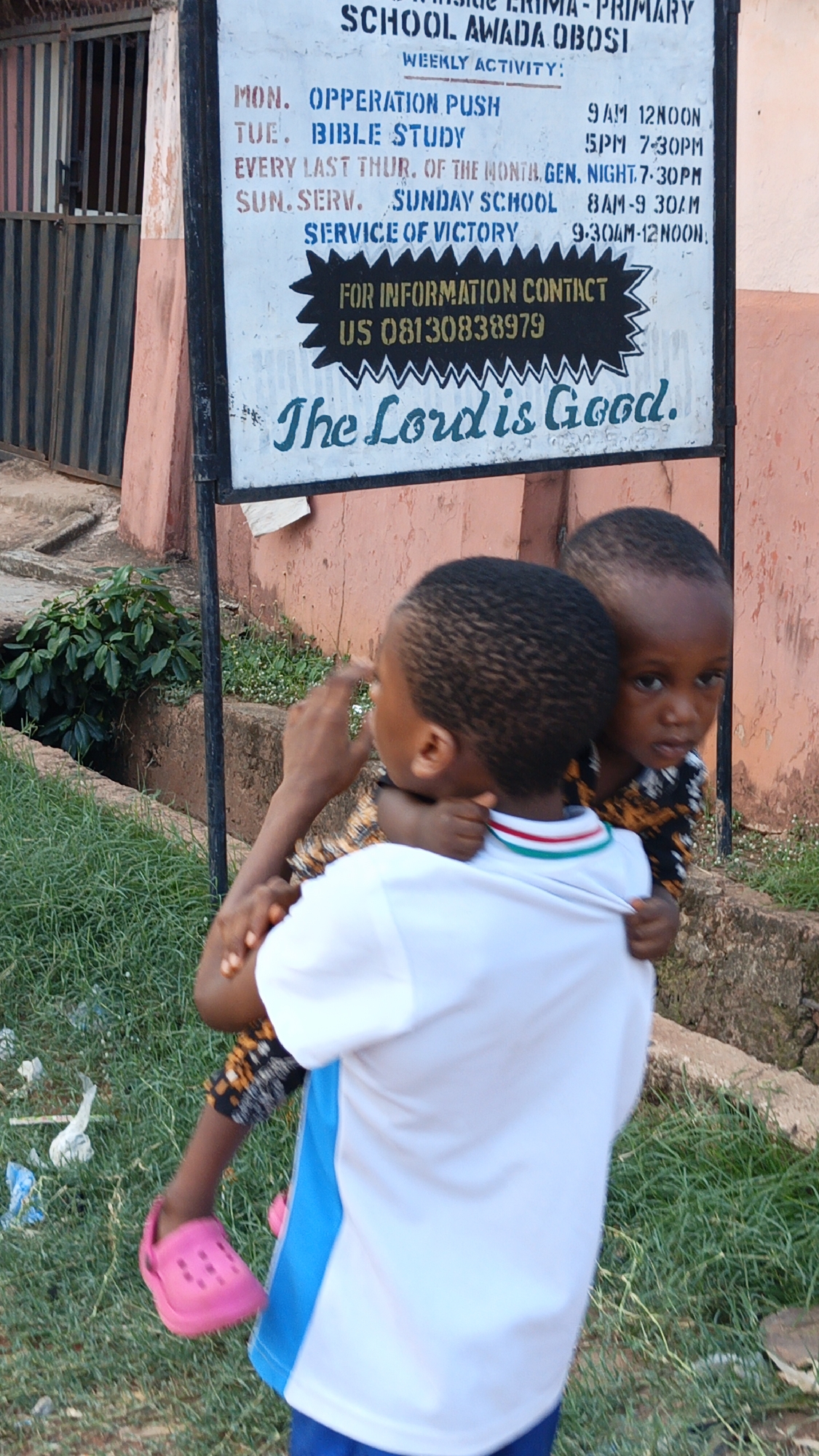In an outdoor scene in Onitsha, Nigeria, two young children are captured in a daytime photograph. An older child, seen from the back, wears a white shirt with blue and red accents, carrying a younger child. The younger child, dressed in a patterned garment and distinctive pink clogs, peeks over the older child's shoulder, gazing directly at the viewer with a neutral expression.
The setting features a concrete wall, overgrown grass, and a visible drainage ditch or gully. In the background, a metal gate, possibly indicating an entrance, is partly visible. To the right, a white signboard displays a "WEEKLY ACTIVITY" schedule, likely belonging to "SCHOOL AWADA OBOSI."
The visible text on the signboard lists:
- "MON. OPPERATION PUSH 9AM 12NOON"
- "TUE. BIBLE STUDY 5PM 7:30PM"
- "EVERY LAST THUR. OF THE MONTH GEN. NIGHT.7-30PM"
- "SUN.SERV. SUNDAY SCHOOL 8AM-9 30AM"
- "SERVICE OF VICTORY 9-30AM-12 NOON"
Below the schedule, it reads: "FOR INFORMATION CONTACT US 08130838979." The bottom of the sign features the phrase, "The Lord is Good." Litter, including plastic scraps, is scattered on the grassy ground near the children.