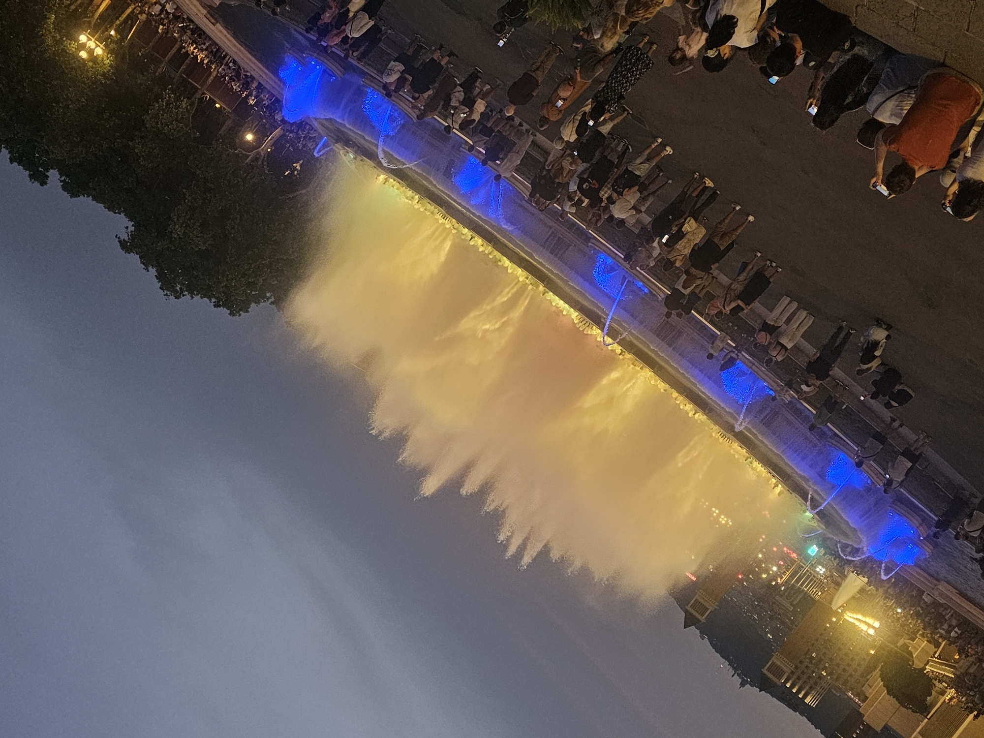 The image depicts the Magic Fountain of Montjuïc in Barcelona, Spain, during an evening performance. The fountain is illuminated with vibrant blue and yellow lights, creating a spectacle of water jets that reach high into the air. A large crowd of people are gathered around the fountain, watching the show. Many are holding up their phones to capture the moment. The surrounding area is dark, with some trees and buildings visible in the background. The sky is a deep blue, indicating that it is nighttime. The scene is dynamic and festive, with the lights and water creating a lively atmosphere.