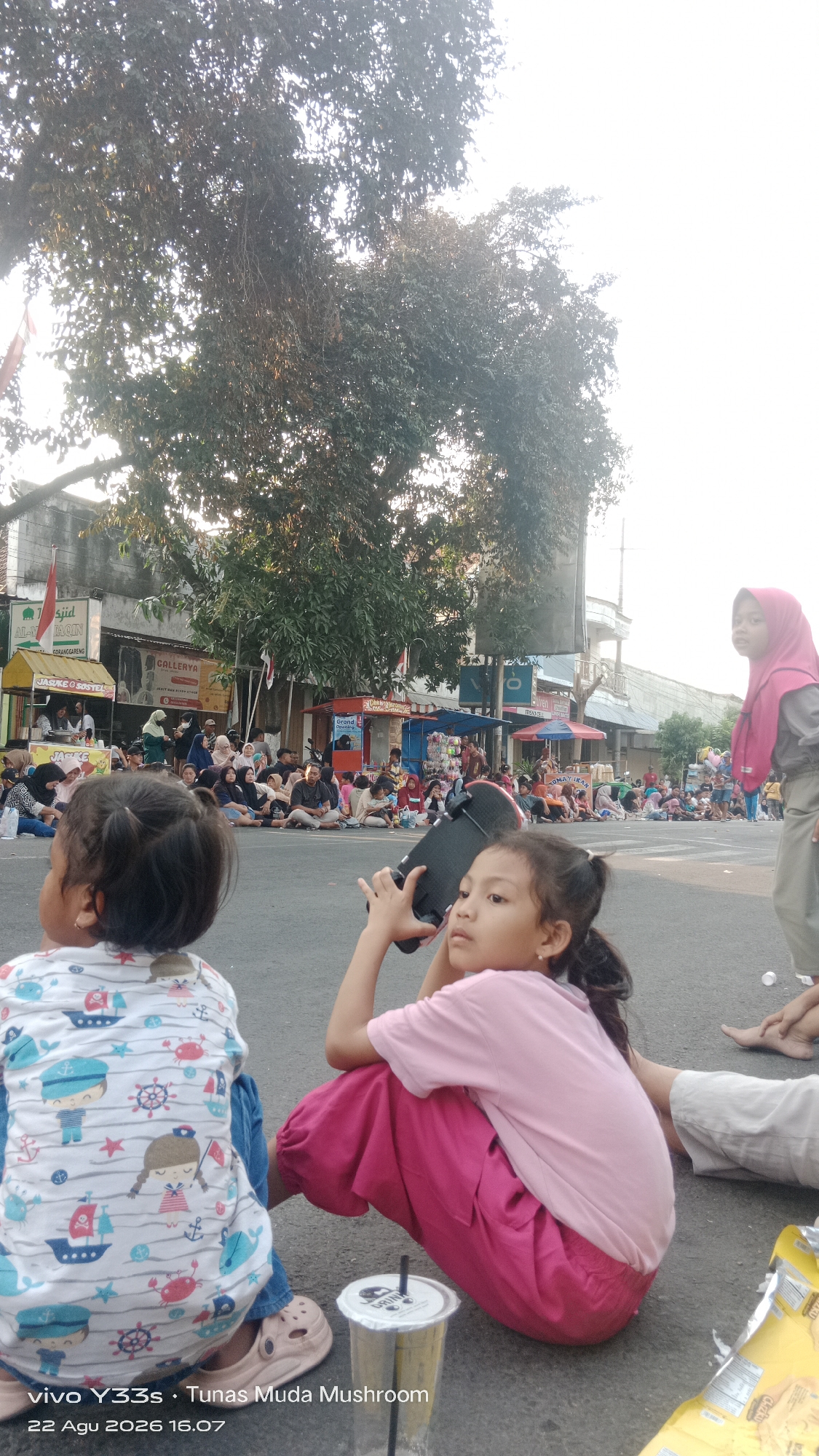 In Genengan, Indonesia, a street scene unfolds with numerous people gathered on the asphalt, suggesting an event or celebration. The foreground features two young girls. The girl on the left, seen from the back, wears a white t-shirt with a nautical-themed print and beige clogs. She is seated on the ground. To her right, a girl in a pink t-shirt and vibrant pink pants is also seated, holding a black object, possibly a camera or a phone, near her face. A beverage cup with a straw sits beside her. In the background, a crowd of individuals, including women wearing hijabs, are seated on the street, facing what appears to be a procession or performance. Several vendor stalls line the street, displaying various goods and signage. Tall trees with dense foliage cast shadows, and buildings with awnings and signs are visible along the street. The sky is bright, indicating daytime. The image captures a community gathering in an outdoor, urban setting.