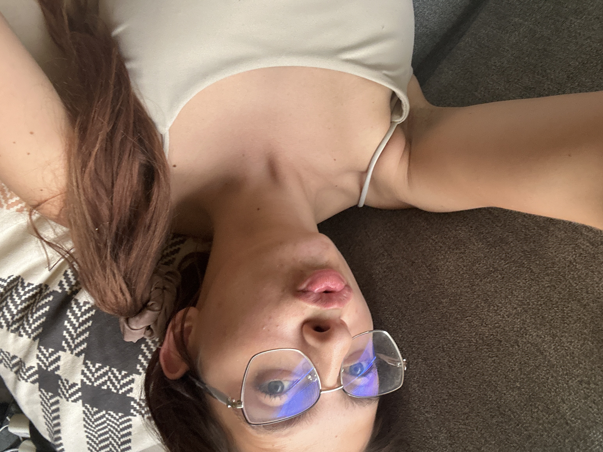 A young woman with long, dark brown hair tied into a ponytail lies on a sofa. She is wearing glasses with silver frames and a light-colored tank top. The sofa is covered with a dark gray textured fabric, and a patterned throw with a black and white geometric design is partially visible in the background. The woman's face is turned towards the camera, and she is puckering her lips. The lighting suggests an indoor setting, possibly a living room. The image was taken in Tryavna, Bulgaria.