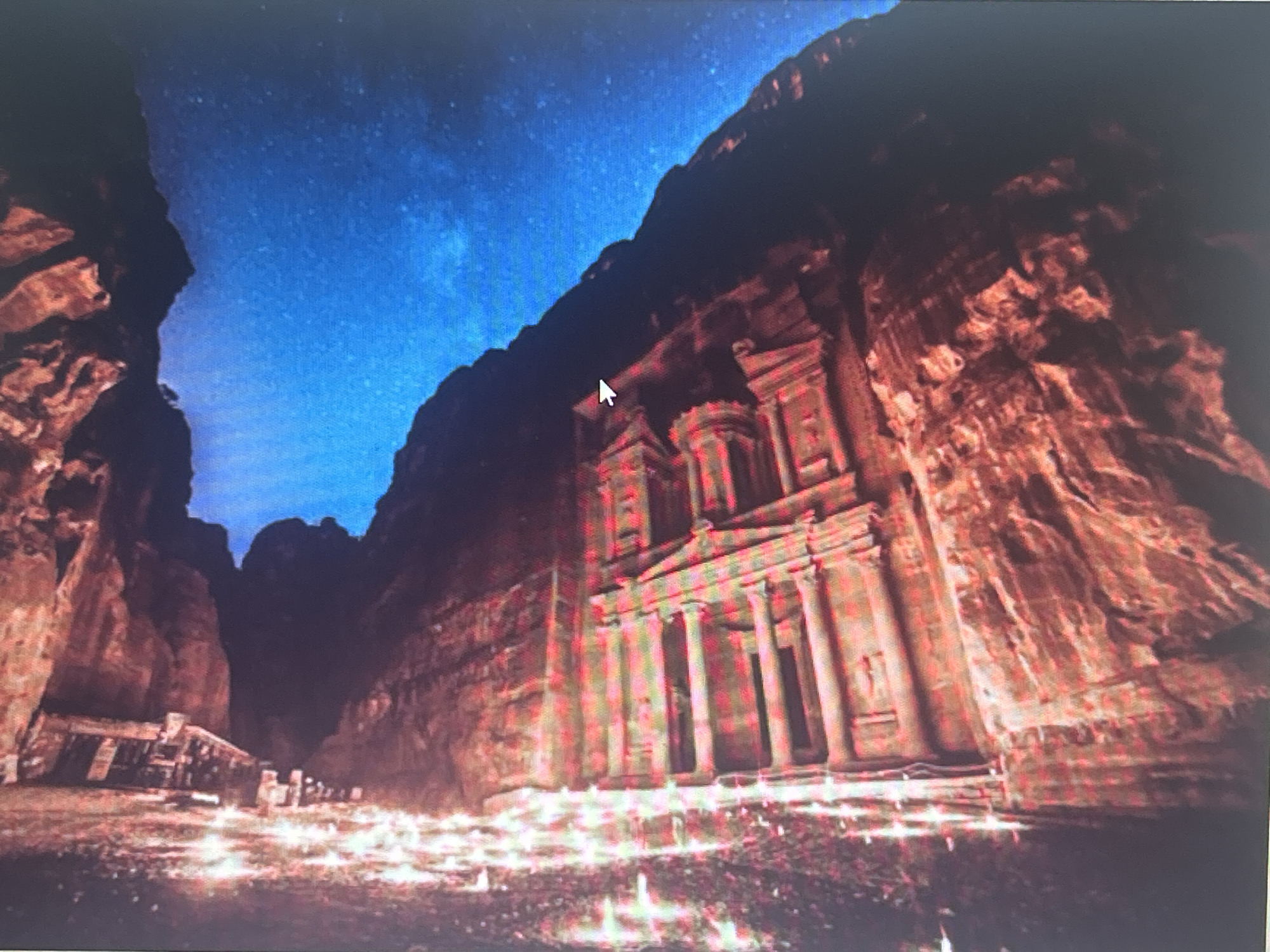 The image depicts Al-Khazneh, also known as The Treasury, a monumental building carved into a sandstone cliff face. The facade features Hellenistic-style columns, pediments, and decorative elements. The structure is illuminated by warm, red-toned lights, highlighting the intricate details of the carvings. The foreground is illuminated by a scattering of small, bright lights, possibly candles or torches, creating a festive or ceremonial atmosphere on the ground. Behind the building, the dark, imposing rock formations of the canyon rise dramatically. Above, the night sky is visible, filled with a multitude of stars, suggesting a clear, dark night. The scene is set in Petra, Jordan, not Sofia, Bulgaria.