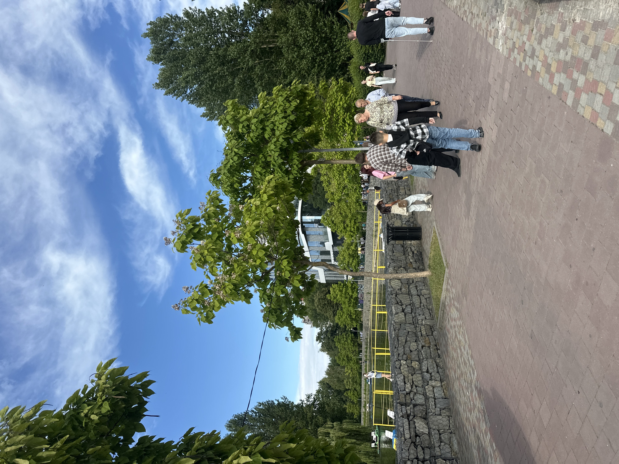 The image captures a sunny day scene in Ternopil, Ukraine, likely at the Ternopil Pond embankment. A paved walkway made of reddish-brown and grey interlocking bricks runs diagonally across the foreground, leading the eye towards the background. To the left of the walkway, a low wall constructed from irregular grey stones lines the edge of a body of water, presumed to be Ternopil Pond. Yellow metal railings extend along this wall. Several paddle boats, two white and one green, are visible on the calm water.
The background reveals a vibrant green park setting with numerous trees. A modern building with a glass facade and white structural elements is situated behind the trees, suggesting a developed urban area adjacent to the park. The sky above is a clear blue with scattered wispy white clouds.
Several people are present in the scene. In the middle ground, a group walks along the brick path. A man with a crutch and light-blue jeans walks towards the right, facing away from the camera. A young boy in a black and white plaid shirt and blue jeans is positioned in the center of the path, also facing away. A woman in a patterned top and dark pants is walking alongside him. Other individuals are dispersed along the path, some walking, some standing. In the distance, near the pond's edge, a person in a white top and blue skirt stands facing the water. The overall atmosphere is leisurely and depicts a public space enjoyed by visitors on a pleasant day.