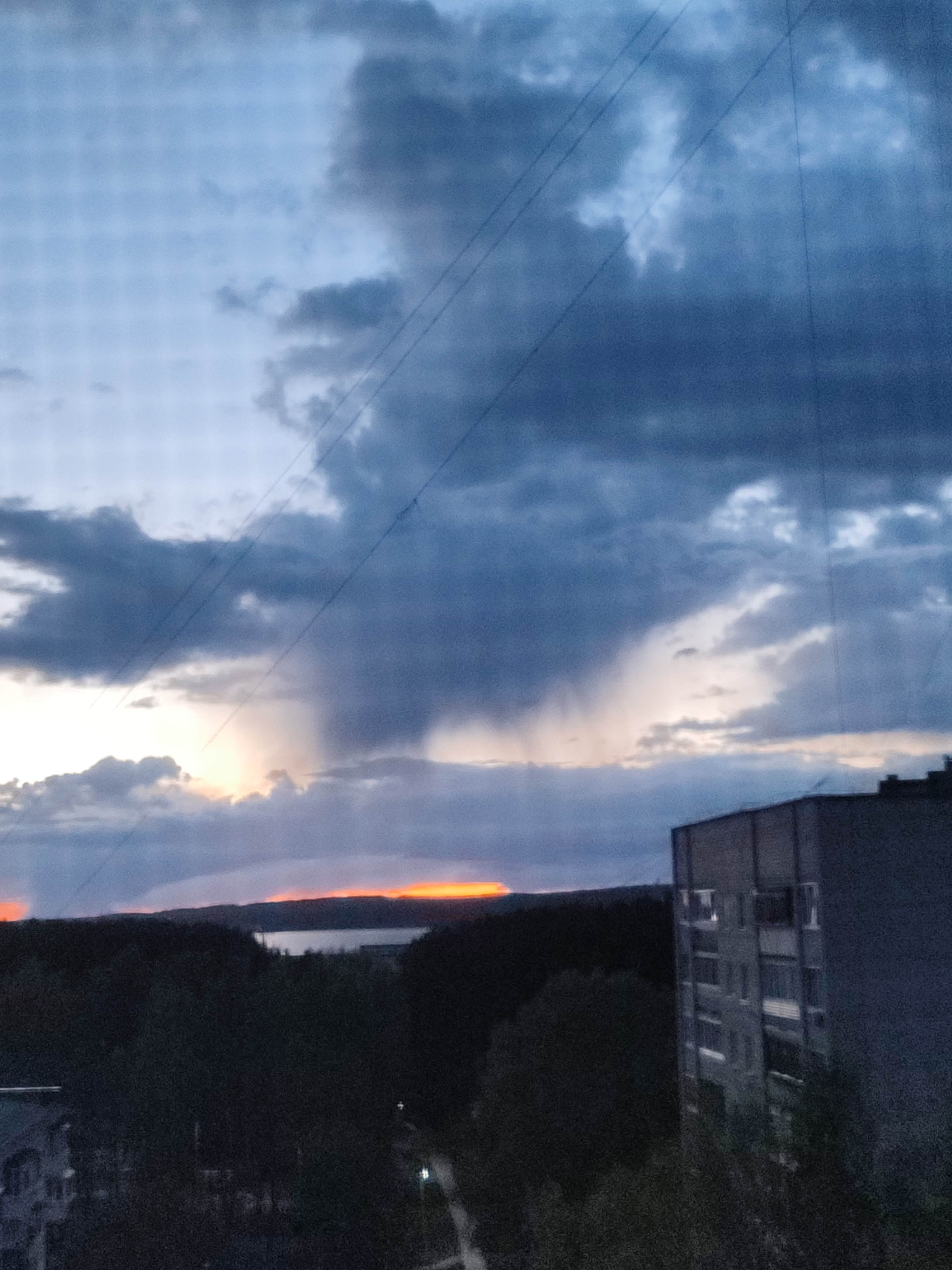 The image depicts a cityscape at dusk or dawn. In the foreground, the edge of an apartment building rises on the right, its concrete facade marked by rows of windows. Below, foliage suggests a wooded area or park interspersed with buildings, which are mostly obscured by the darkness. In the middle distance, a narrow body of water is visible, reflecting a band of fiery orange light from the horizon. The sky is dominated by dark, ominous clouds with areas of rain visible, adding to the turbulent and uncertain atmosphere. Power lines run across the sky. The location seems to be a residential area on the edge of a city, bordering a natural landscape. The overall impression is one of impending storm or a scene after a storm, with the sun peeking through the clouds.
