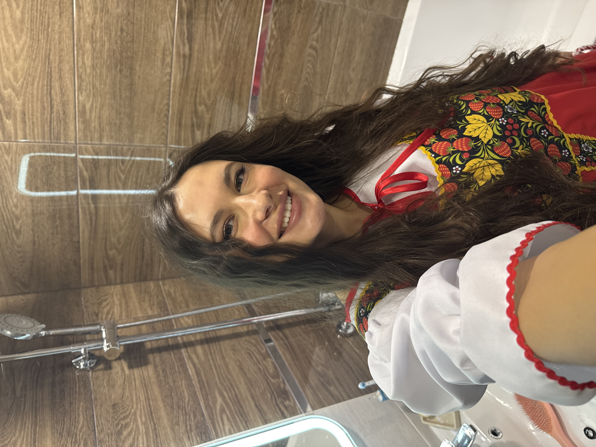 A woman is smiling in a bathroom, possibly in Unknown, Unknown, taking a selfie while wearing a traditional Russian costume. The costume features a red and black floral pattern with yellow accents, a white blouse with red trim, and red ribbons. The bathroom has wood-paneled walls, a chrome showerhead, and a white sink with a chrome faucet. A pink hairbrush is also visible on the sink.