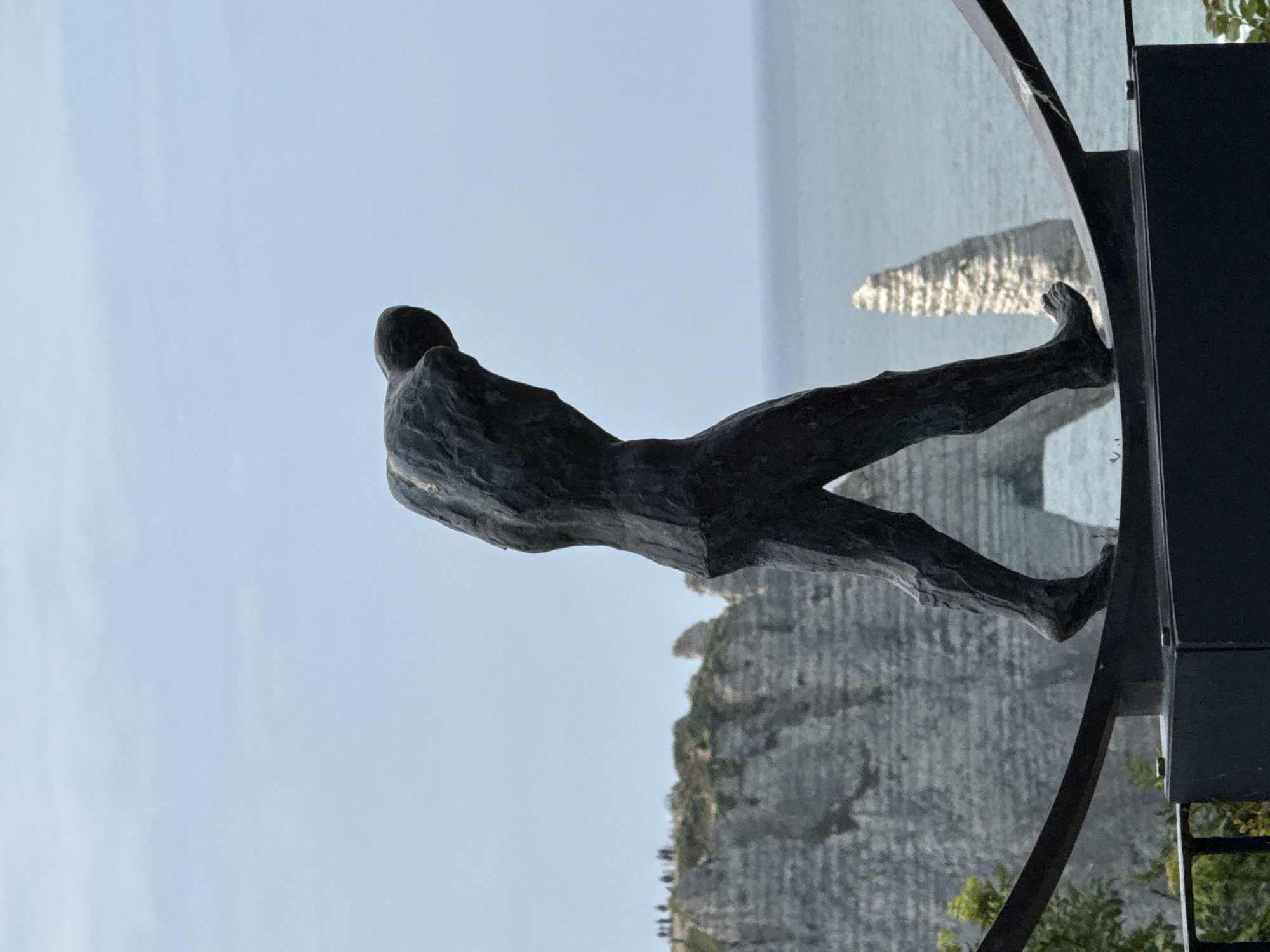 A dark, textured, and abstract sculpture of a human figure, appearing to be a man, stands prominently in the foreground, gazing out towards a vast seascape. The sculpture is crafted from a rough, dark material, possibly weathered bronze or stone, with a somewhat blocky and angular form. It is positioned as if leaning forward slightly, observing the distant horizon.
In the background, a calm, expansive body of water, likely the sea or ocean, stretches under a clear, pale blue sky. Dramatic white cliffs, characteristic of coastal formations, rise sharply from the water on the right side of the image. These cliffs display vertical striations and rugged textures, with sparse vegetation visible in some areas. A distinctive sea stack or rock formation stands isolated in the water near the cliffs.
The scene is illuminated by bright daylight, suggesting a clear and sunny day, likely late morning or afternoon. A dark, curvilinear metal structure, possibly a railing or part of the sculpture's installation, is visible along the right edge, framing the view. The overall impression is one of quiet contemplation amidst a stunning natural coastal landscape. Given the contextual information, the location is identifiable as Étretat, France, known for its iconic chalk cliffs and arches. No text is visible in the image.