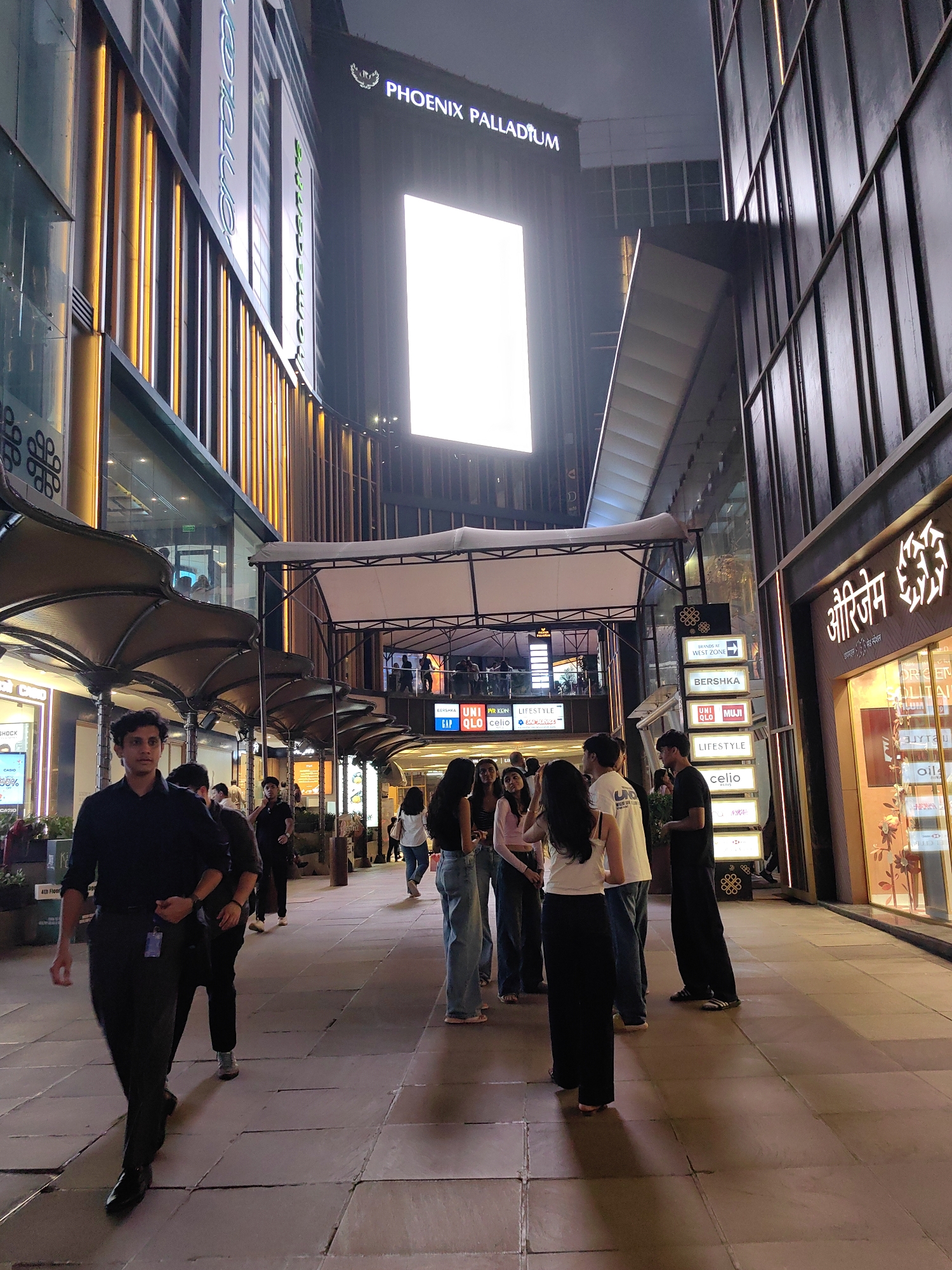 The image captures a nighttime scene at Phoenix Palladium, a prominent shopping mall in Mumbai, India. The architecture of the mall features modern, sleek designs with extensive use of glass and metal. A large, illuminated screen is visible high up on one of the buildings, displaying bright white light. Vertical golden lights accentuate the facade of a building on the left.
A walkway paved with square tiles forms the central part of the scene, with a canopy of umbrella-like structures providing shade. People are scattered across the walkway, some walking and others standing in small groups. On the right side of the walkway, a shopfront with illuminated signage in what appears to be Hindi is visible, along with storefronts for well-known brands like Zara, Celio, and lifestyle stores. Several people are seen walking towards the viewer, with one man in a black shirt and dark trousers in the foreground. Another group of people, mostly young adults, are gathered further down the walkway. The overall atmosphere suggests a bustling evening at a popular urban commercial and entertainment destination.