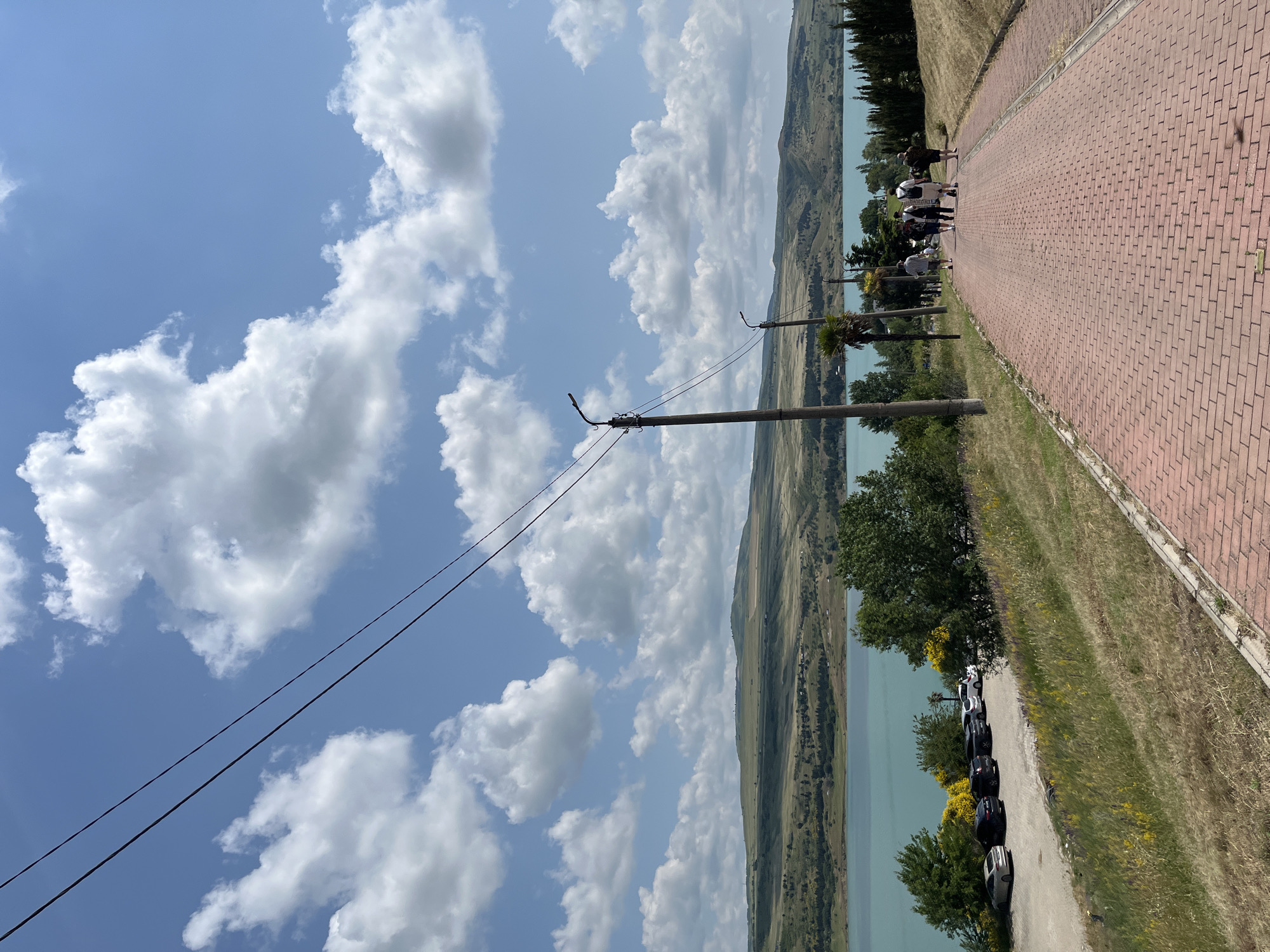 The image depicts a scenic view of the Tbilisi Sea, a reservoir located in Tbilisi, Georgia. The foreground features a red brick pathway leading uphill, bordered by dry grass and scattered yellow wildflowers. Several individuals, appearing to be tourists, are walking up the pathway in the distance. To the left of the pathway, a parking lot is visible, with a line of cars parked on a gravel surface. Beyond the parking lot, lush green trees and shrubs separate the area from the calm, turquoise waters of the Tbilisi Sea. The background showcases rolling, verdant hills under a bright blue sky dotted with fluffy white clouds. Power lines stretch across the upper portion of the frame, supported by utility poles along the pathway. The overall scene conveys a sense of outdoor recreation and natural beauty.