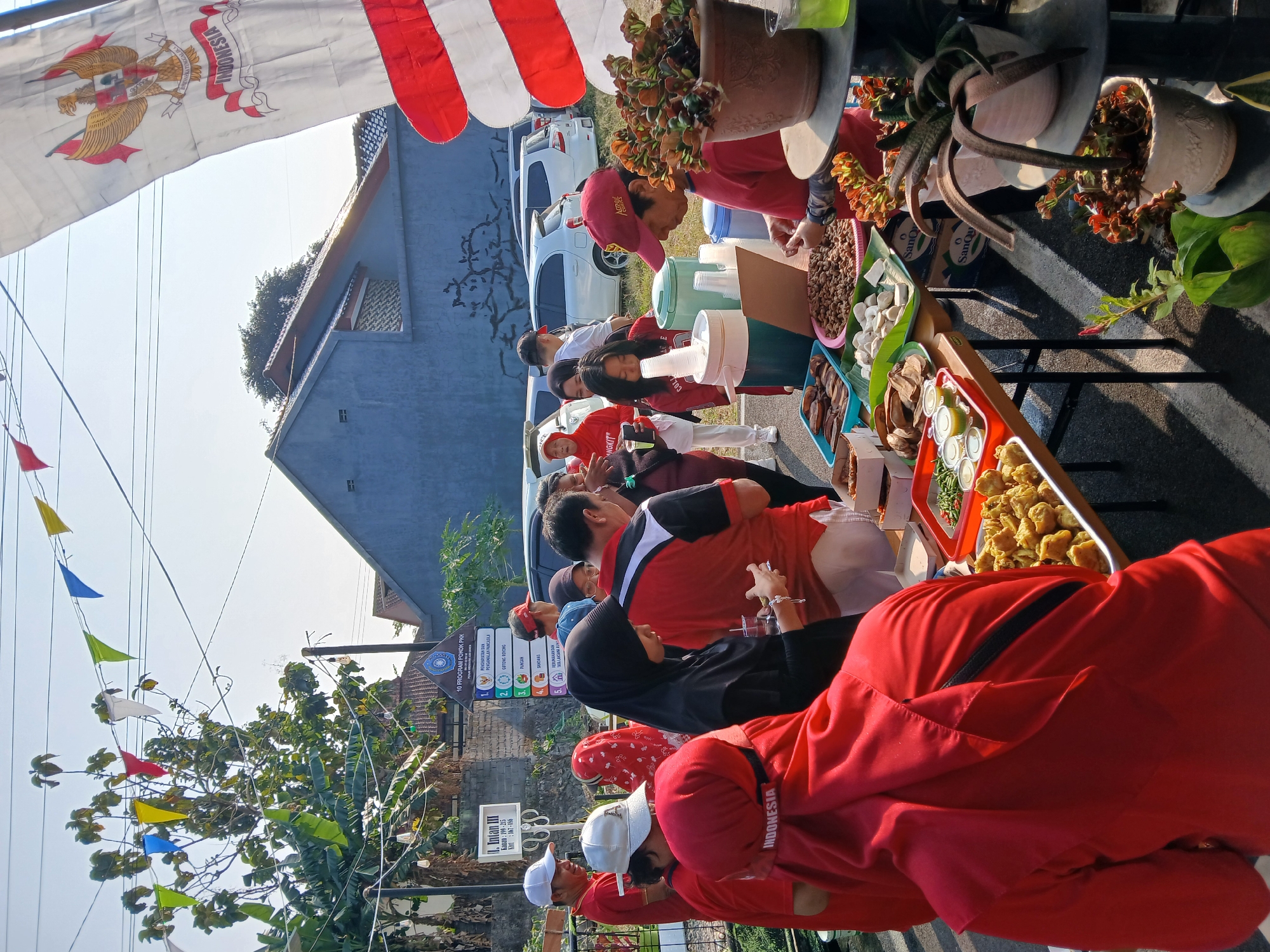 This image depicts an outdoor event or market scene, likely in Ambarawa, Indonesia, given the Indonesian flag and context. The foreground is dominated by a table laden with various food items, including fried snacks, meatballs, and nuts, presented in trays and containers. Several people, dressed in casual attire with some wearing red shirts or headscarves, are gathered around the table, appearing to be customers or vendors. One person in a red shirt and cap is actively serving or preparing food. Behind the table, a line of cars is parked. Decorative triangular flags in various colors are strung across overhead wires, and a large Indonesian flag with Garuda Pancasila is visible on the right. The background shows a building with a dark blue facade and a residential structure with a tiled roof. The overall atmosphere suggests a community gathering or a local food stall setup.