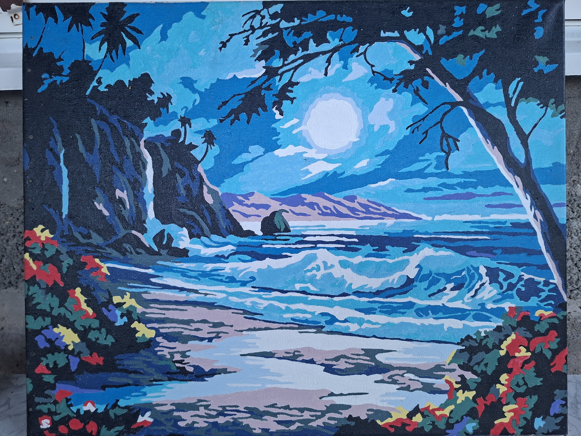 This is a painting of a nighttime beach scene. A large, bright moon hangs in a blue sky filled with clouds. Palm trees silhouette against the sky on the left side of the painting, and a large tree branch hangs into the frame from the top right. The ocean is depicted with waves crashing on the shore. In the foreground, there are bushes with red, yellow, and blue flowers. There are no people or animals in the painting. The painting is done in a blocky, stylized manner, characteristic of a paint-by-numbers kit. The painting is on canvas and is framed. There is no information to suggest this scene is located in Gorkovskiy, Russia; the imagery suggests a tropical or coastal environment.