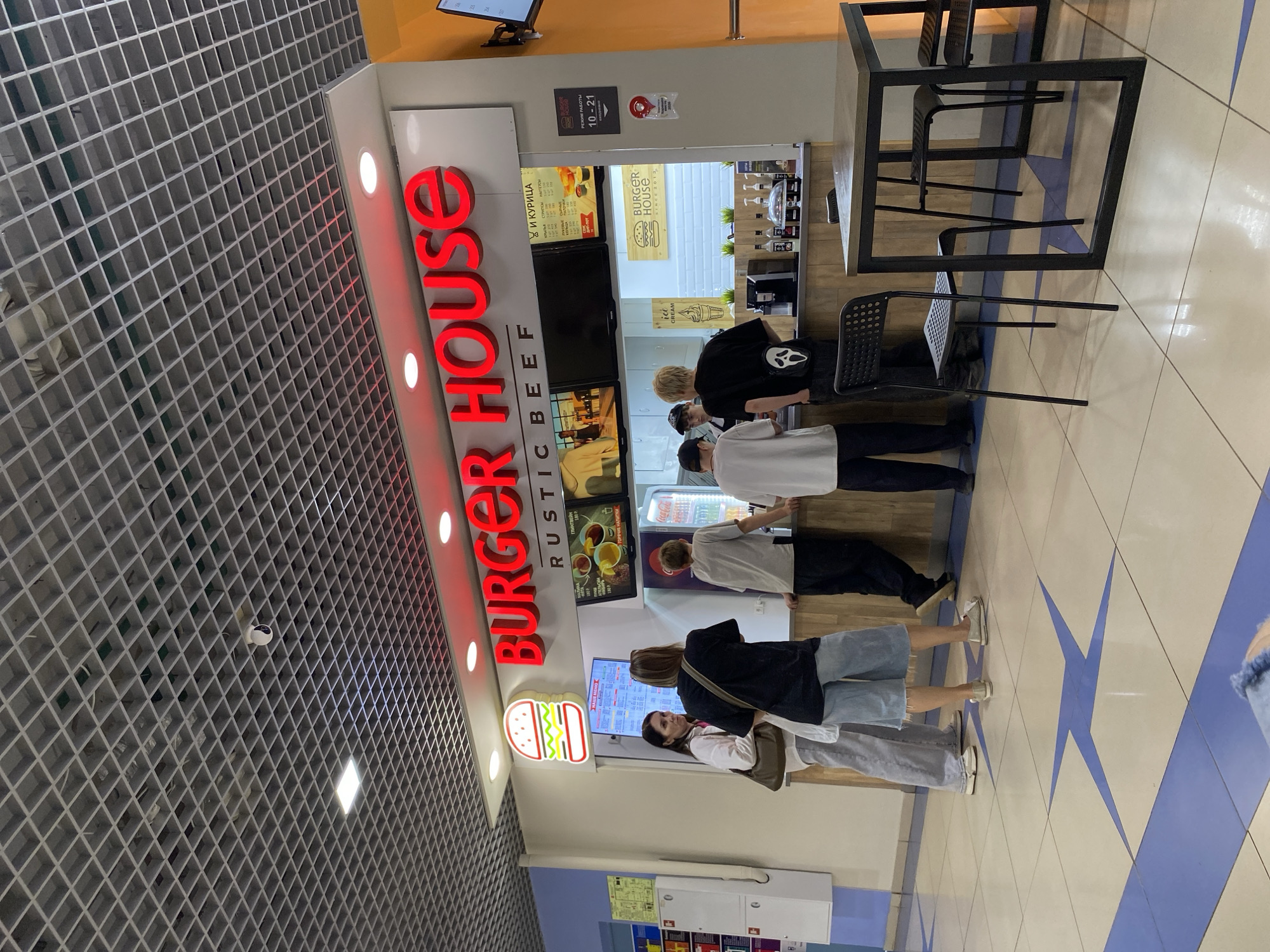 This media file shows a group of people standing in line at a fast-food restaurant called "BURGER HOUSE RUSTIC BEEF". The restaurant appears to be located in a mall or similar indoor public space, indicated by the tiled floor and grid-patterned ceiling with a security camera. The restaurant's signage is prominent, featuring large red letters spelling out "BURGER HOUSE" and a cartoon burger logo.
The people in the image are of various ages and are dressed casually. They are waiting to order food, with some looking at menus displayed on screens or posters. One person is wearing a black t-shirt with a white ghost-like face on it.
Visible text on the signage includes "BURGER HOUSE", "RUSTIC BEEF", and in Russian, "АПЕЛЬСИН" (Orange), "ГОРЯЧИЕ НАЛИТКИ" (Hot Drinks), and "И КУРИЦА" (And Chicken). There is also a sign indicating "РЕЖИМ РАБОТЫ 10-21", which translates to "Operating Hours 10-21". The presence of an "ice CREAM" sign suggests ice cream is also sold.
The lighting in the scene is bright, consistent with an indoor commercial environment. The overall mood appears neutral, with people engaged in the common activity of purchasing food. The setting is clean and well-lit. The blue star pattern on the floor is a notable decorative detail. The image is taken from a slightly elevated angle, looking down at the scene.