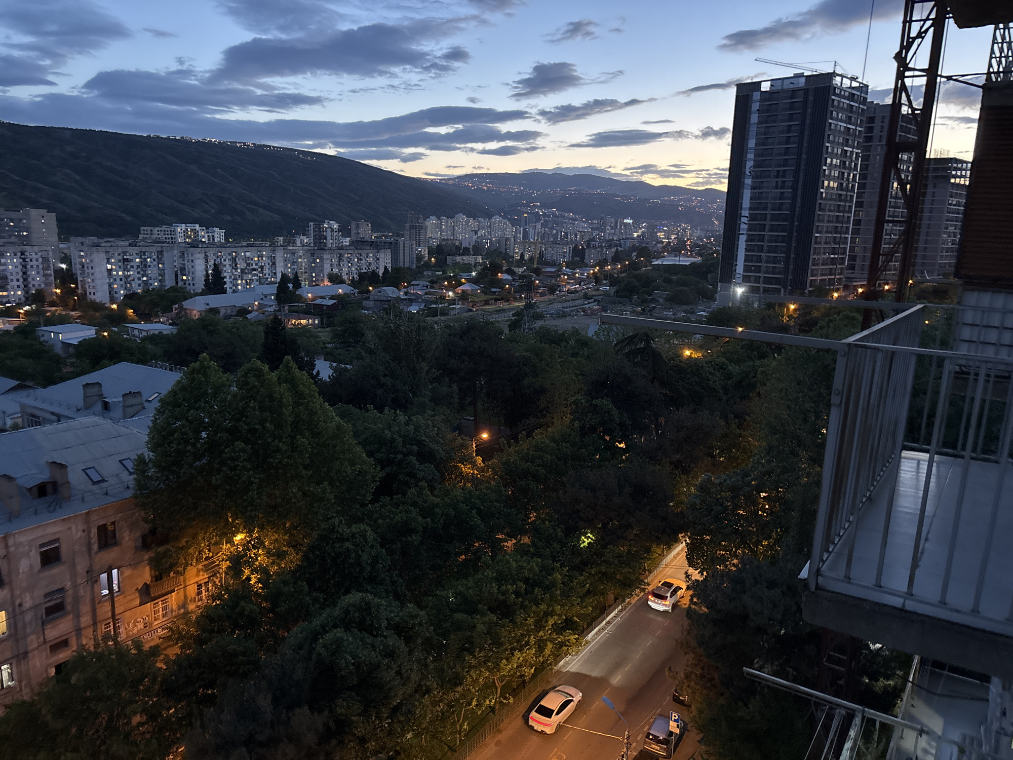 The image depicts a nighttime cityscape of Tbilisi, Georgia, viewed from an elevated perspective, likely a balcony. In the foreground, there's a metal balcony railing. Below, a street is illuminated by streetlights, with several cars parked along the side and a few moving. The scene is heavily populated with trees obscuring much of the lower view. Beyond the trees, a cityscape extends towards a distant mountain range silhouetted against the twilight sky. Numerous buildings are visible, including high-rise apartment complexes under construction, and older residential buildings. The sky is a mix of blues and grays with streaks of orange from the fading sunlight. The overall atmosphere is one of urban tranquility in the evening.