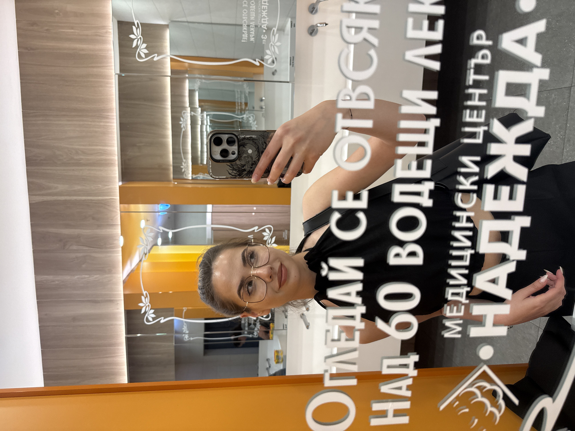 A woman is taking a selfie in a mirror. The mirror has the name "Nadezhda Medical Center" and other information written on it in Bulgarian, including "Look from every angle," "More than 60 leading doctors," and what appears to be "Medical Center." The location is in Sofia, Bulgaria. The woman is wearing glasses and a black top. The phone she is holding is dark in color. The background is a wooden panel and what appears to be a display of some kind.