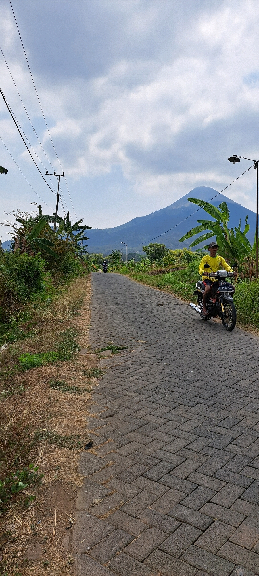 In Sumber Suko, Indonesia, a person wearing a yellow shirt and a cap rides a motorcycle down a paved road. The road is constructed from interlocking grey bricks laid in a herringbone pattern and is lined with dry grass on the left and green vegetation on the right. In the background, a large mountain dominates the skyline under a cloudy sky. Banana trees are visible to the right of the road, and power lines stretch across the sky. Another motorcycle is seen in the distance further down the road.