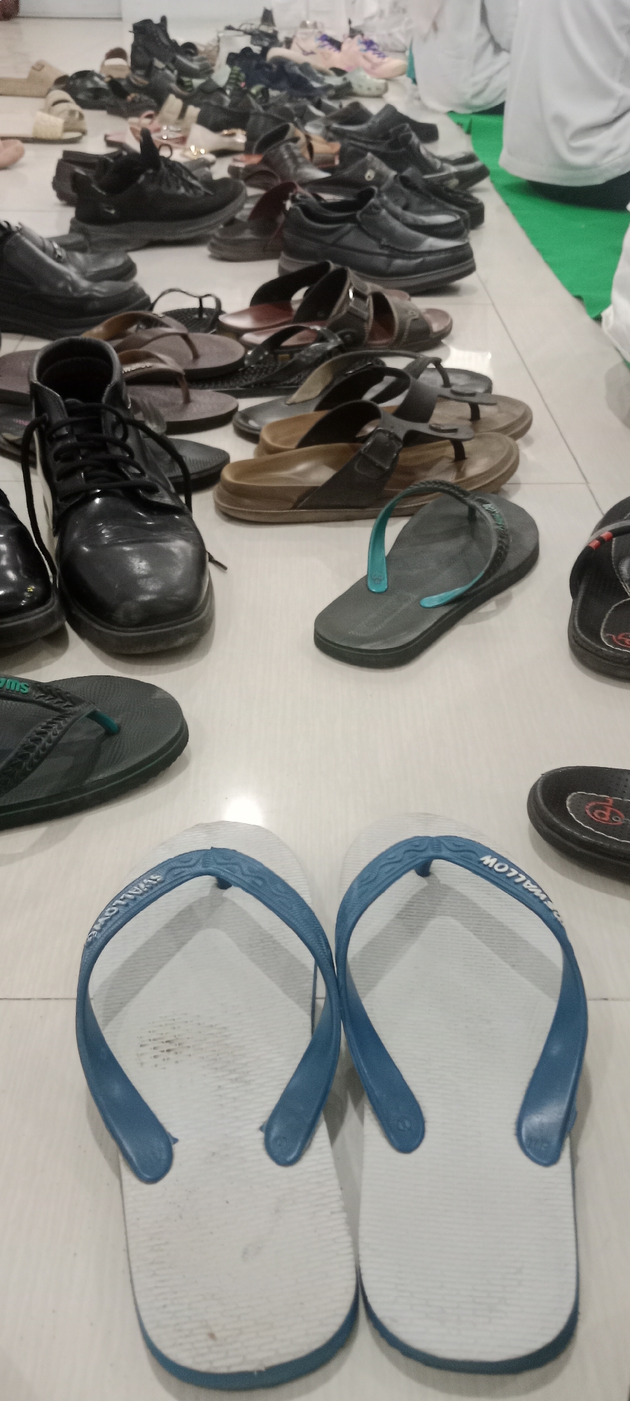 The image displays a collection of footwear, primarily men's shoes and sandals, arranged on a tiled floor. In the foreground, a pair of white flip-flops with blue straps are prominently featured. Behind them, a variety of other shoes are visible, including black leather shoes, boots, and numerous pairs of sandals and flip-flops in different colors and styles. Some individuals in white clothing are partially visible in the background, seated on a green mat, suggesting a religious gathering or event. The scene appears to be indoors, likely a public space where shoes are removed before entering, common in religious sites or during specific ceremonies. The overall impression is one of an orderly arrangement of footwear, possibly at the entrance of a mosque or a similar venue in Tuban, Indonesia.