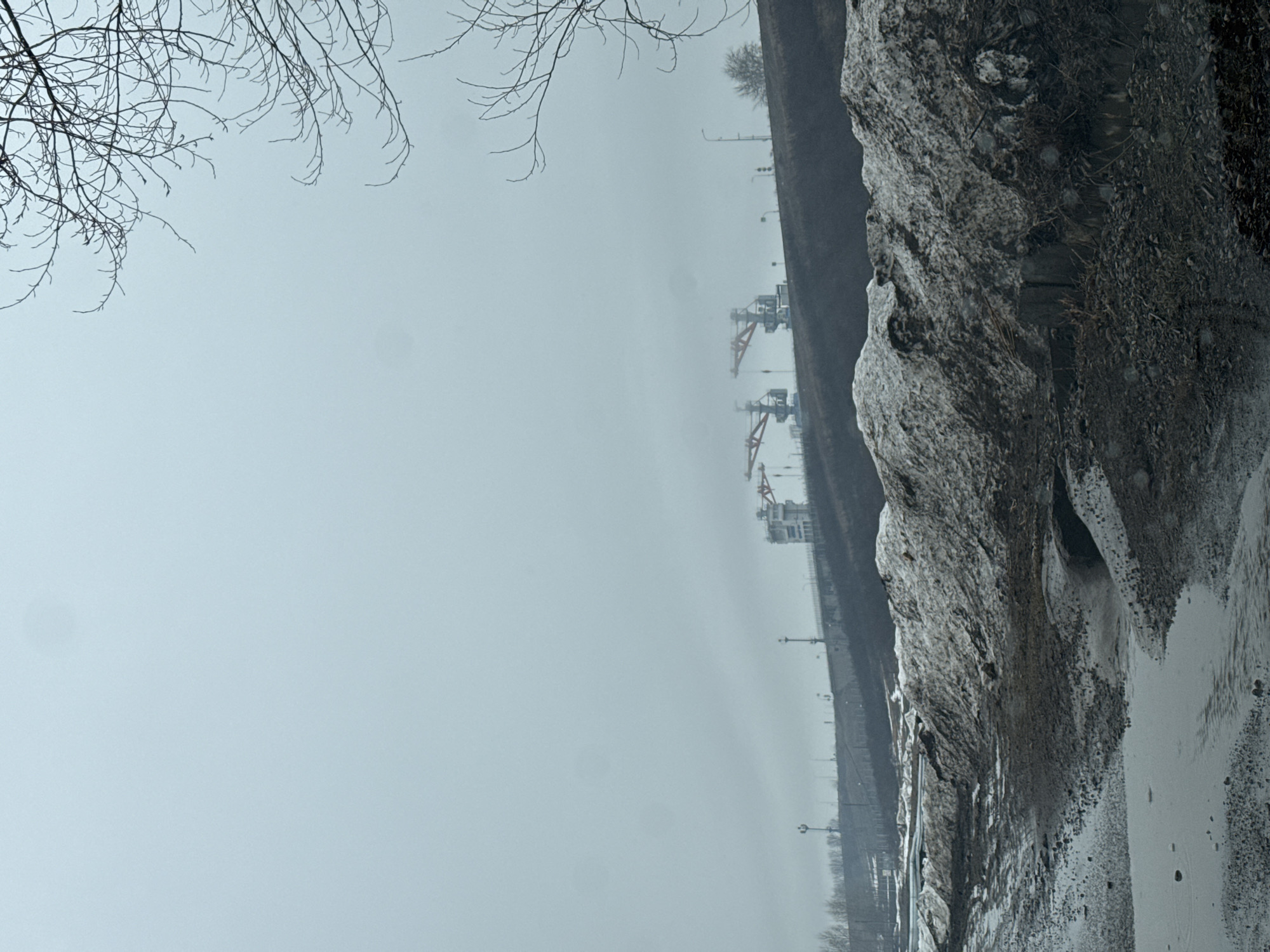 The image shows a winter scene near the Kama River in Chaikovsky, Russia. In the distance, partially obscured by fog, are industrial cranes likely associated with the Kama shipyard. Along the right side of the image is a snow-covered embankment or riverbank. The foreground includes the bare branches of a tree. The sky is overcast and gray.