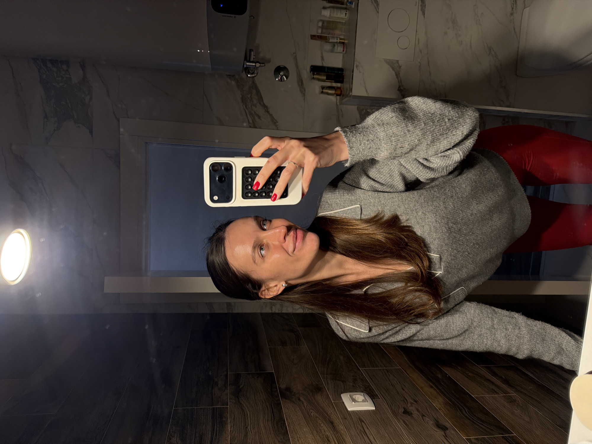The image depicts a female subject capturing a mirror selfie within a bathroom setting. The subject, a woman with long dark brown hair and light blue eyes, appears to be in her 20s or 30s. She is dressed in a grey knitted V-neck sweater with white trim and bright red, glossy-textured pants. A silver hoop earring is visible in her left ear, and a silver ring is present on the ring finger of her left hand. Her fingernails are painted red. She holds a white smartphone, encased in a white protective cover featuring a grid of black circular indentations, with her left hand. Her gaze is directed to her right, away from the phone's camera.
The immediate environment visible in the mirror reflection includes a ceiling-mounted circular recessed light fixture. The left wall displays dark brown, horizontally arranged wood-grain patterned tiles, on which a white electrical outlet switch is mounted. The right wall area features light-colored marble-patterned tiles. Reflecting in the mirror are a portion of a white water heater/boiler with a blue indicator light, a white toilet bowl, and a white flush plate with two circular buttons installed above the toilet. A narrow shelf holding several small bottles and containers is also visible on the right. A section of a white counter or sink edge is present at the bottom of the frame.
The image contains no specific visual data to ascertain a precise location within the city of Budva, Montenegro.